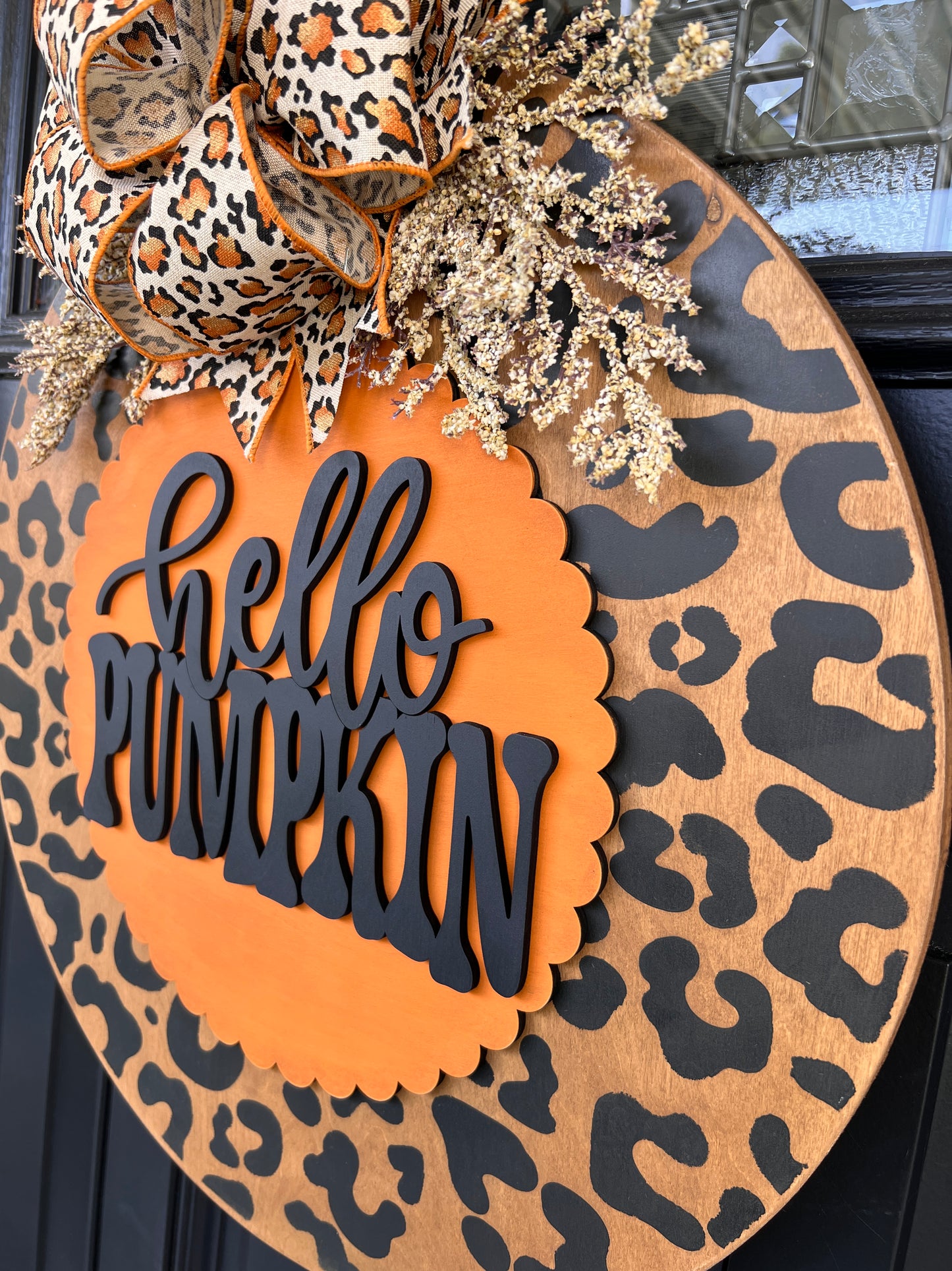 Hello Pumpkin leopard print fall door hanger with layered wood lettering, designer bow, seasonal greenery, and woodgrain animal print background displayed on a front door