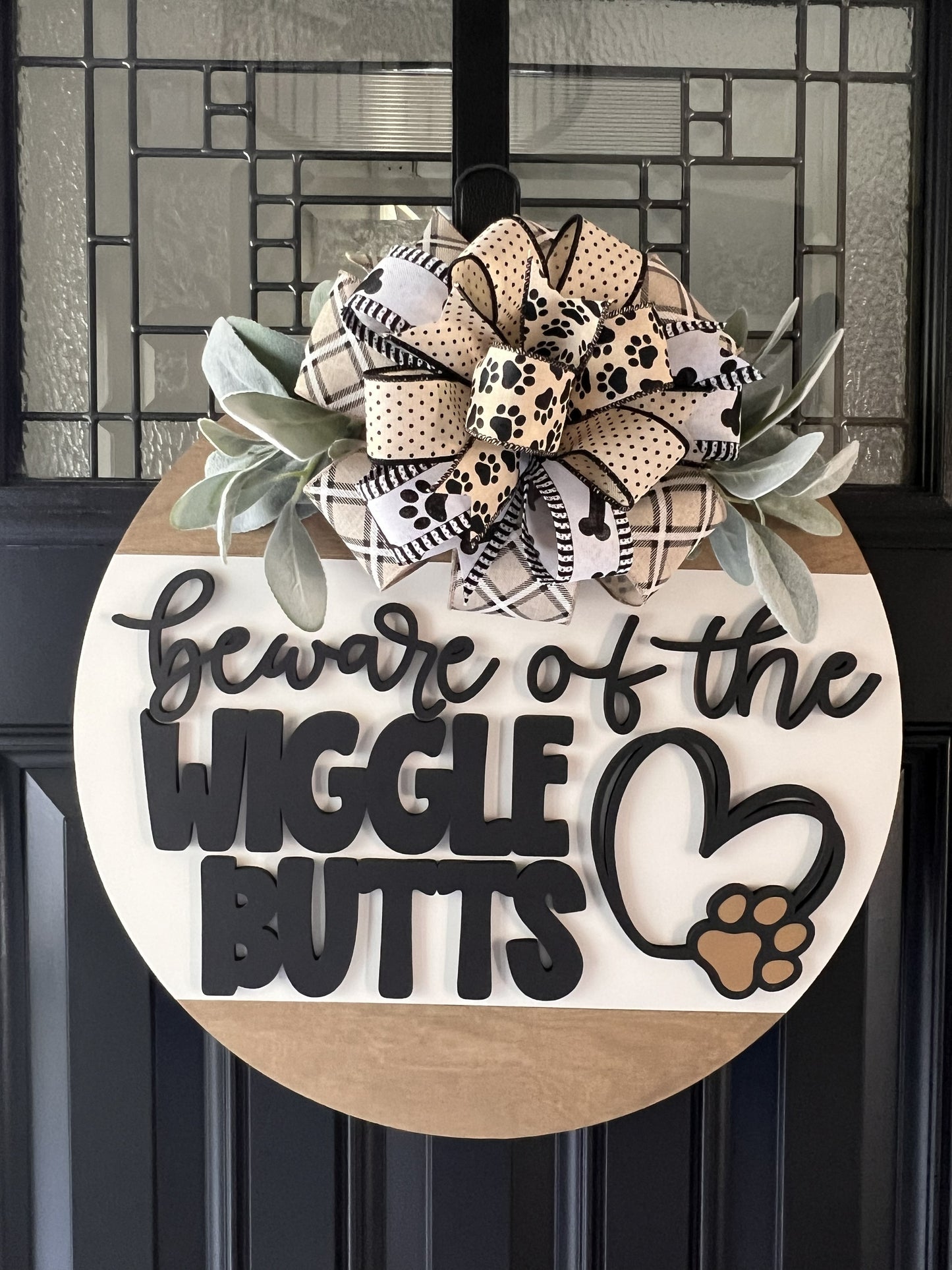 Decorative door hanger with a pawprint bow and text 'Beware of the Wiggle Butts' with bow and greenery on a black door.