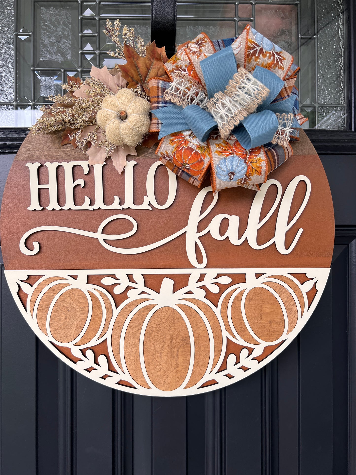 Hello Fall pumpkin door hanger with layered wood lettering, decorative pumpkin cutouts, designer bow, seasonal greenery, and autumn accents displayed on a front door