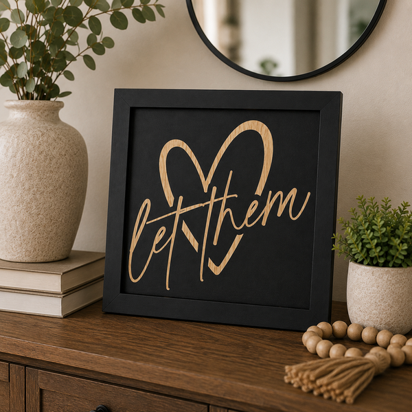 A 10x10 square black-framed wall sign with a natural wood heart and the phrase 'let them' in cursive script, styled on a wooden console table with a vase, books, and a mirror in the background.