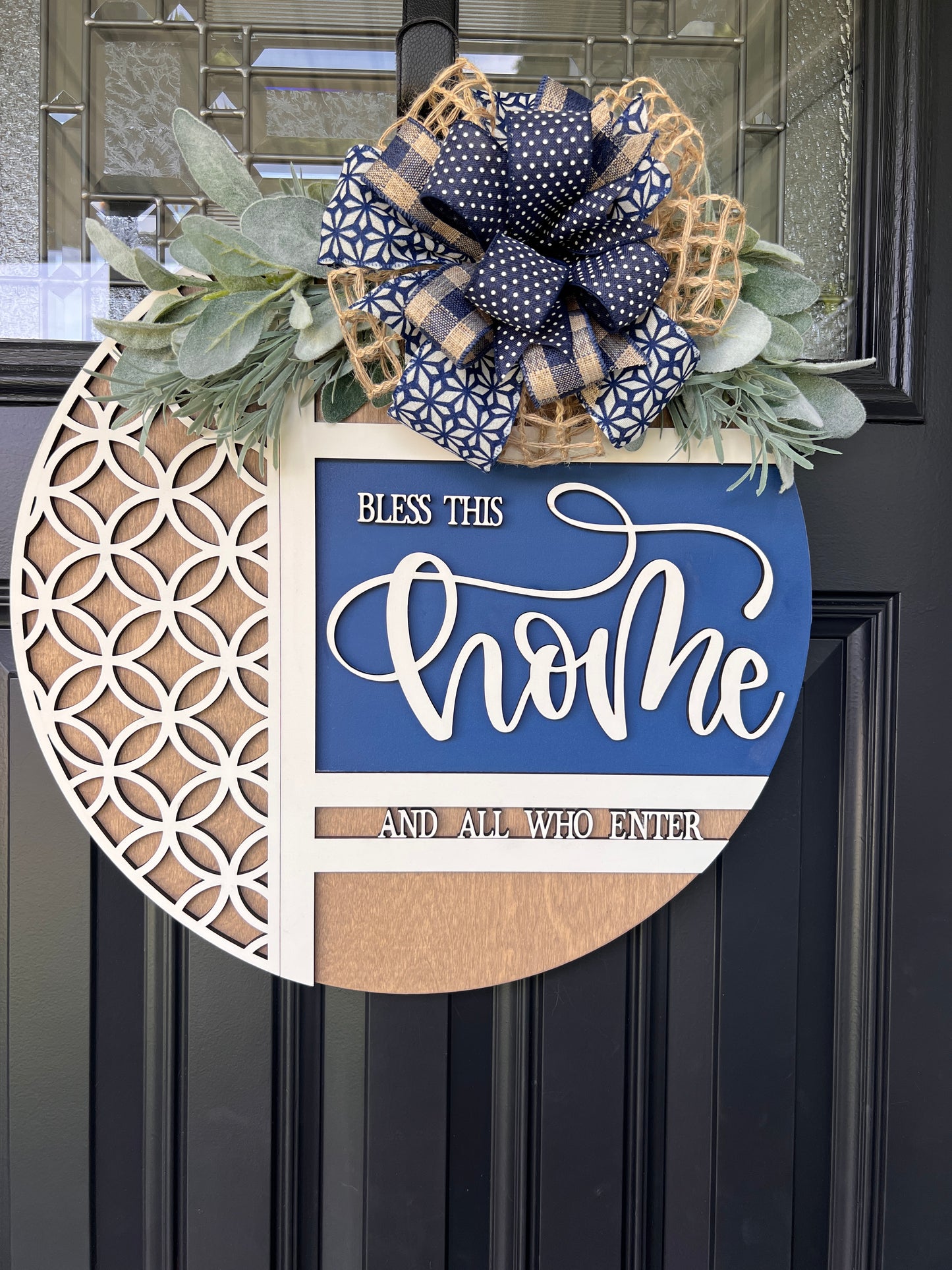 Navy blue and natural wood Bless This Home door hanger with layered 3D lettering, geometric overlay design, greenery accents, and designer bow displayed on a black front door.