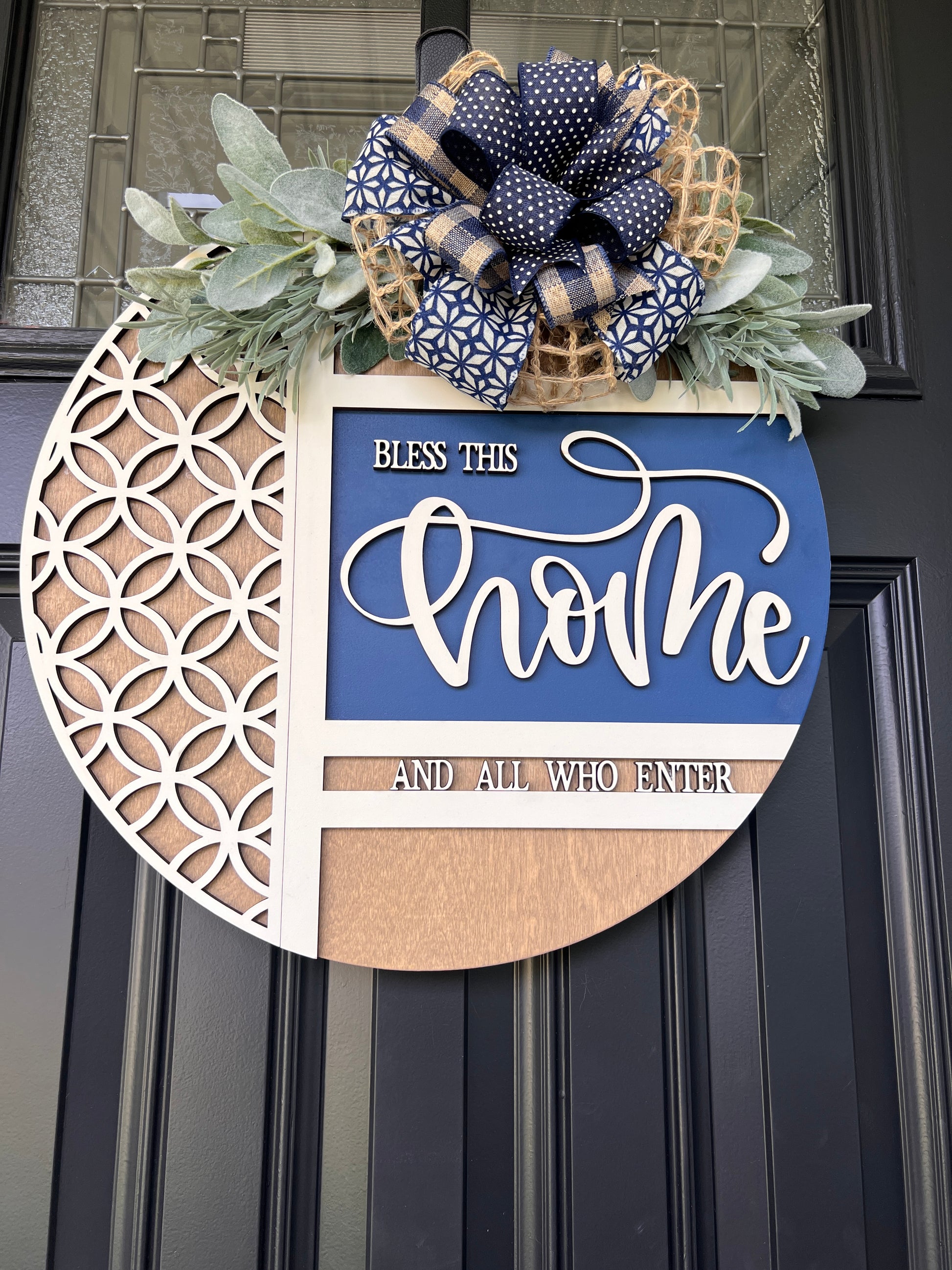 Navy blue and natural wood Bless This Home door hanger with layered 3D lettering, geometric overlay design, greenery accents, and designer bow displayed on a black front door.