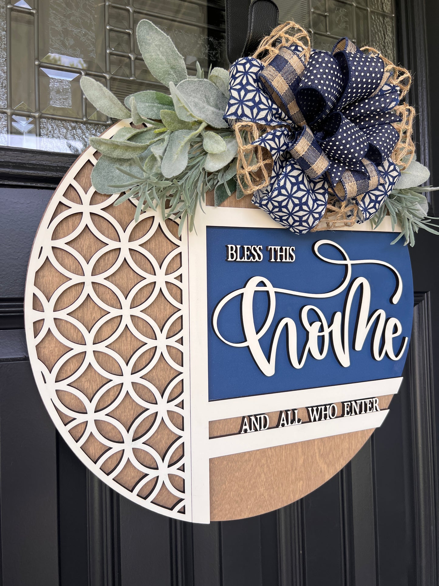 Navy blue and natural wood Bless This Home door hanger with layered 3D lettering, geometric overlay design, greenery accents, and designer bow displayed on a black front door.