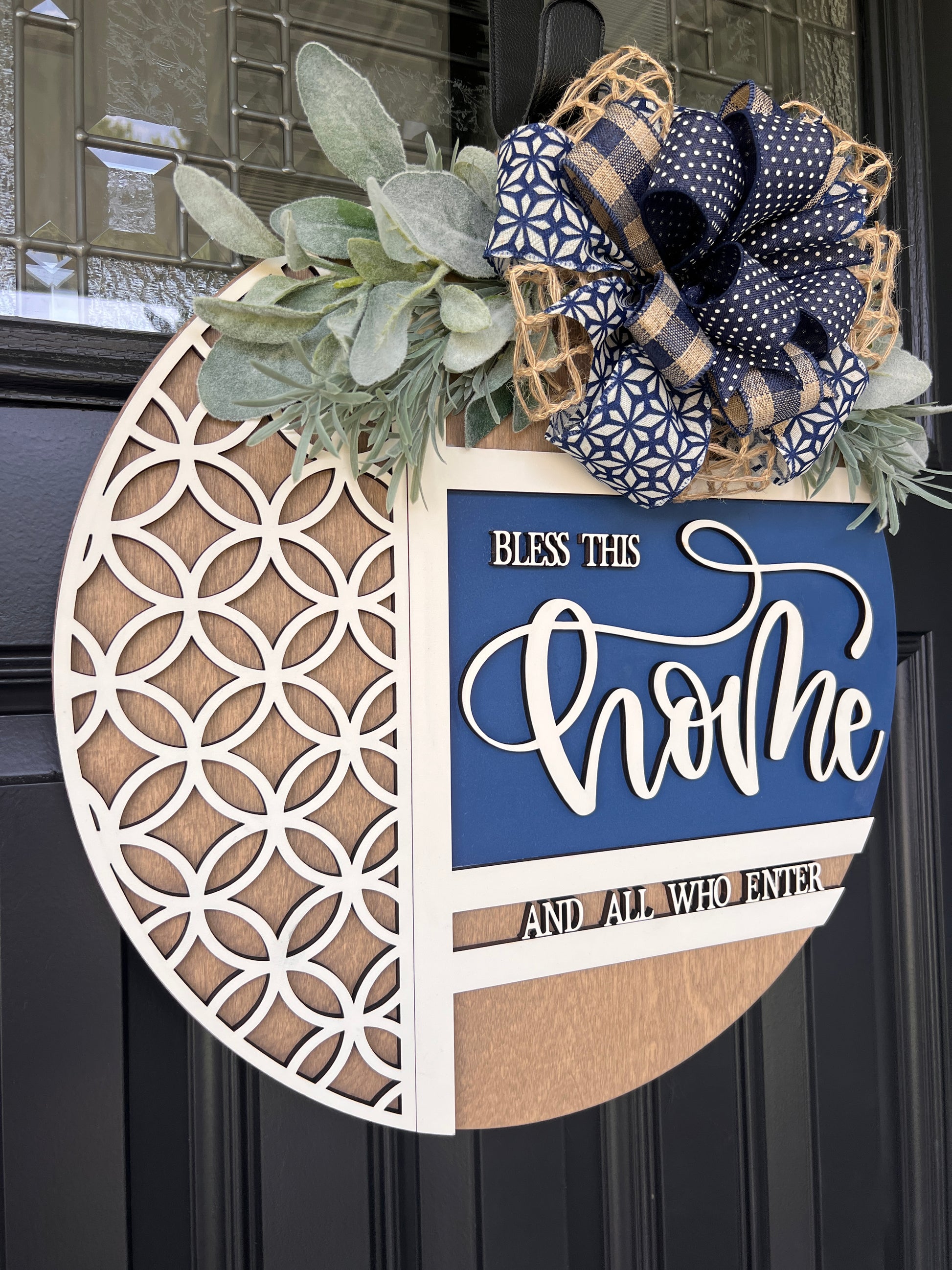 Navy blue and natural wood Bless This Home door hanger with layered 3D lettering, geometric overlay design, greenery accents, and designer bow displayed on a black front door.