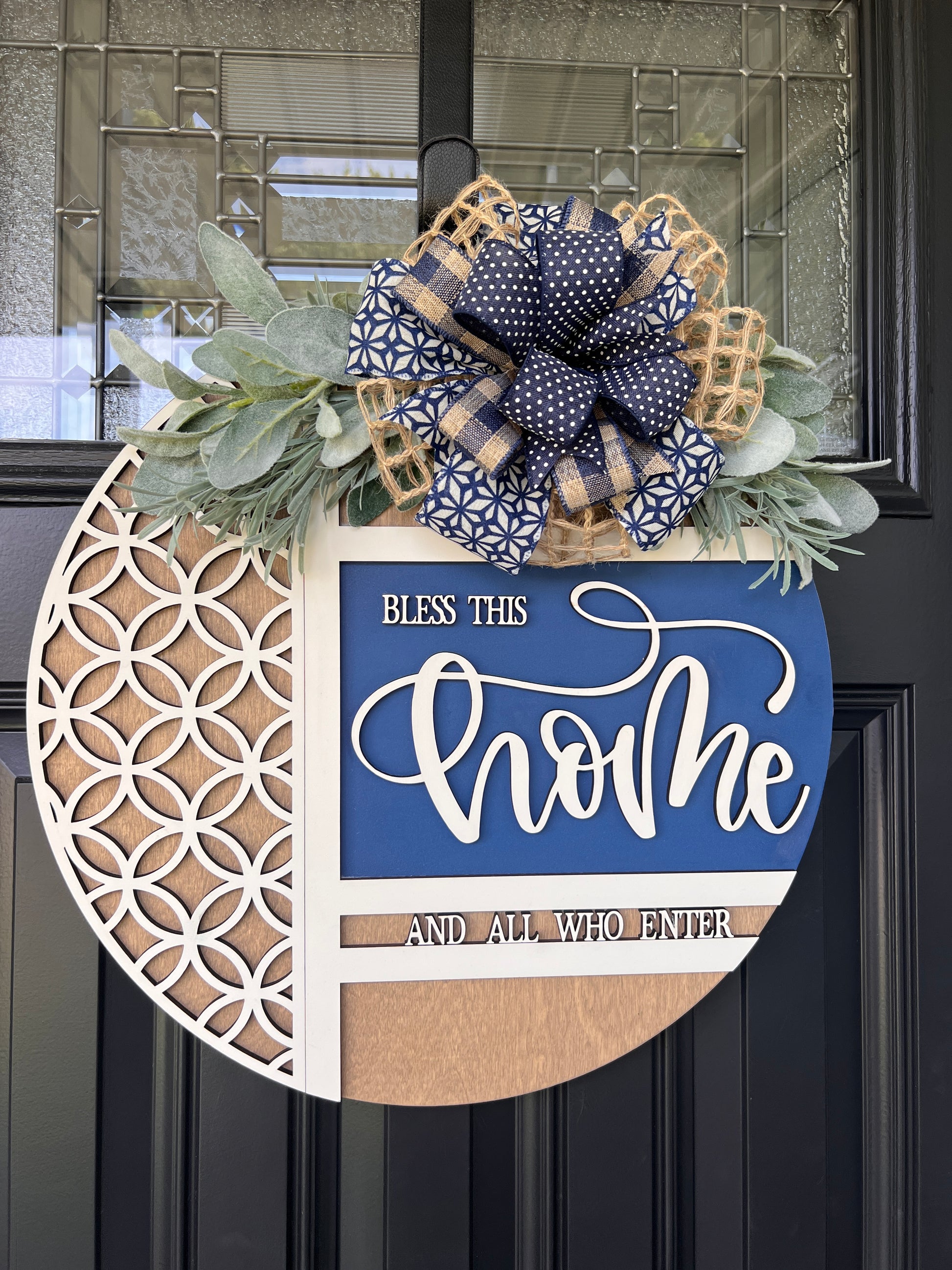 Navy blue and natural wood Bless This Home door hanger with layered 3D lettering, geometric overlay design, greenery accents, and designer bow displayed on a black front door.