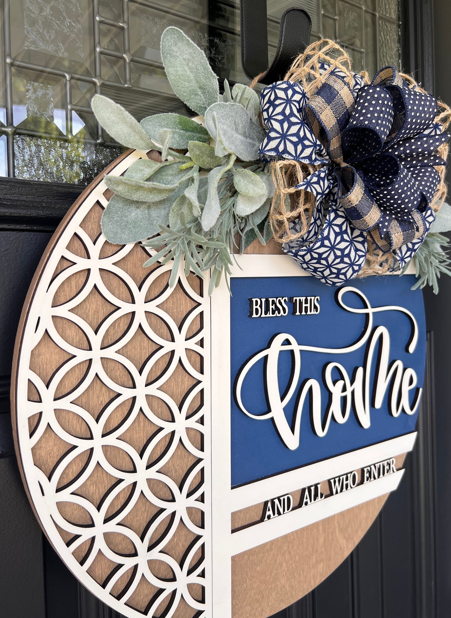 Navy blue and natural wood Bless This Home door hanger with layered 3D lettering, geometric overlay design, greenery accents, and designer bow displayed on a black front door.