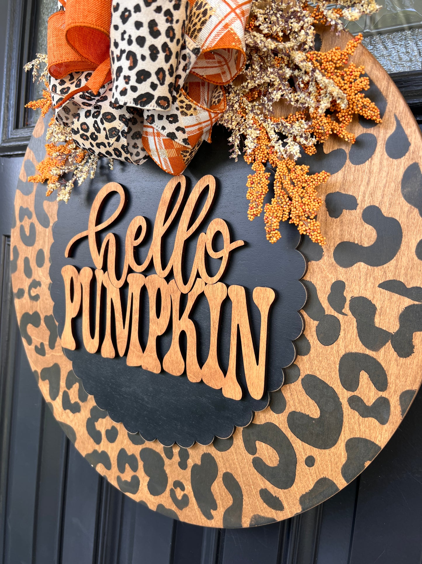 Hello Pumpkin leopard print fall door hanger with layered wood lettering, designer bow, seasonal greenery, and woodgrain animal print background displayed on a front door