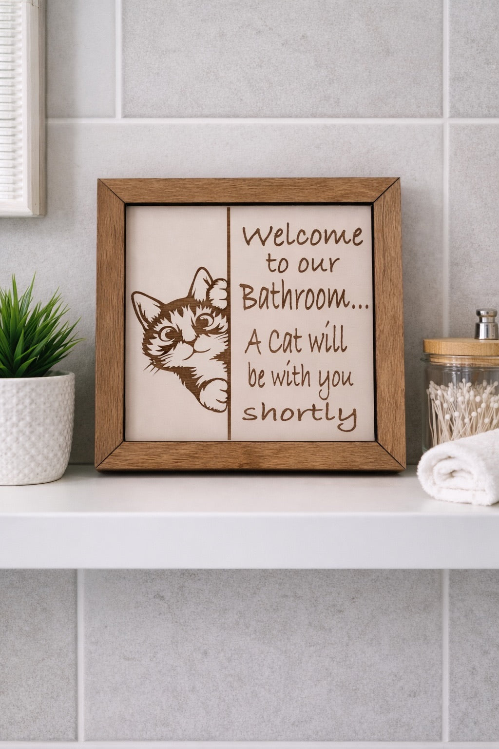 Welcome to Our Bathroom A Cat Will Be With You Shortly engraved birch wood sign with peeking cat graphic, styled on a white bathroom shelf with green plant and cotton swab jar