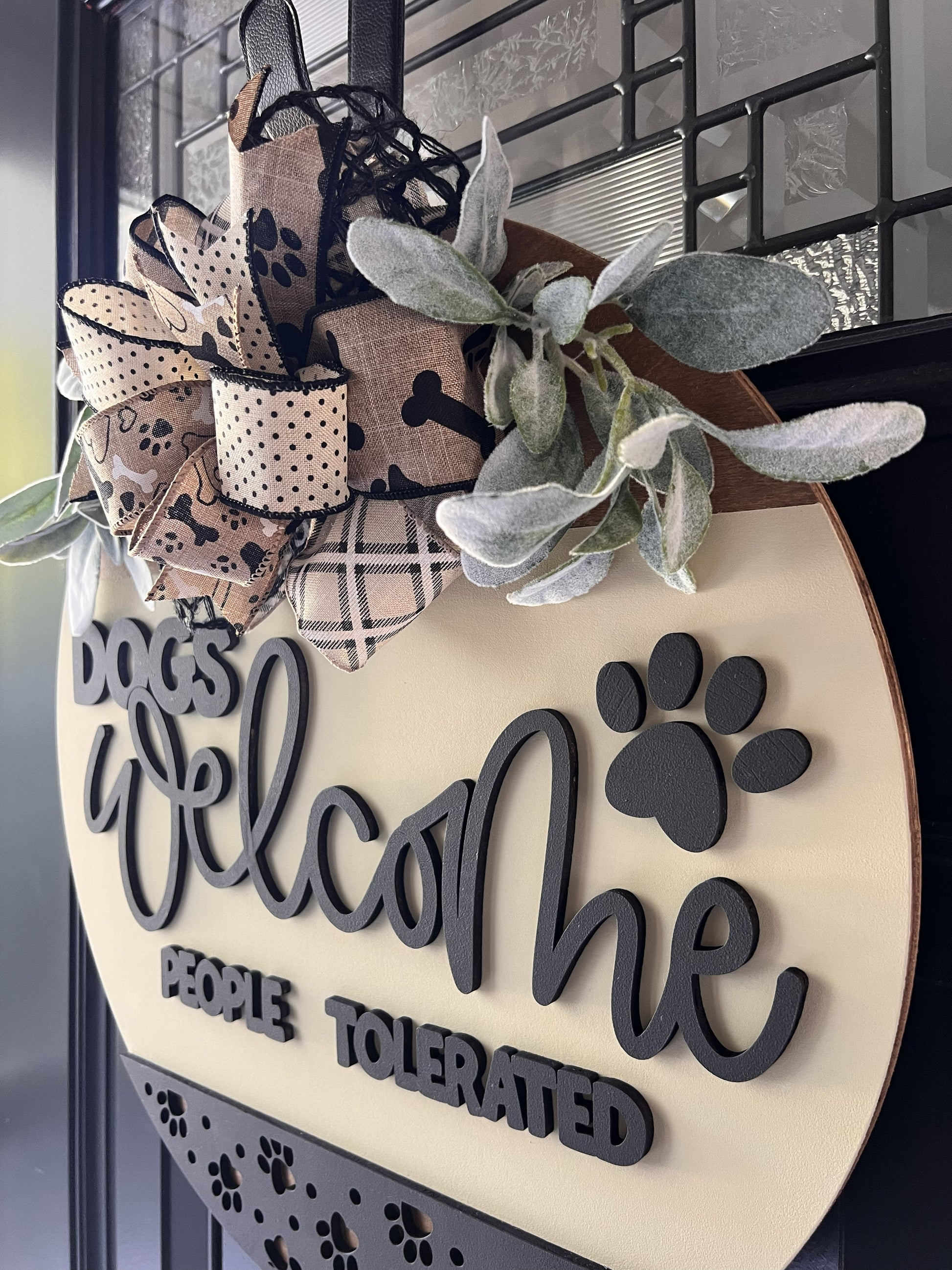 “Dogs Welcome People Tolerated wooden door hanger with layered lettering, paw print accents and decorative bow on front door”