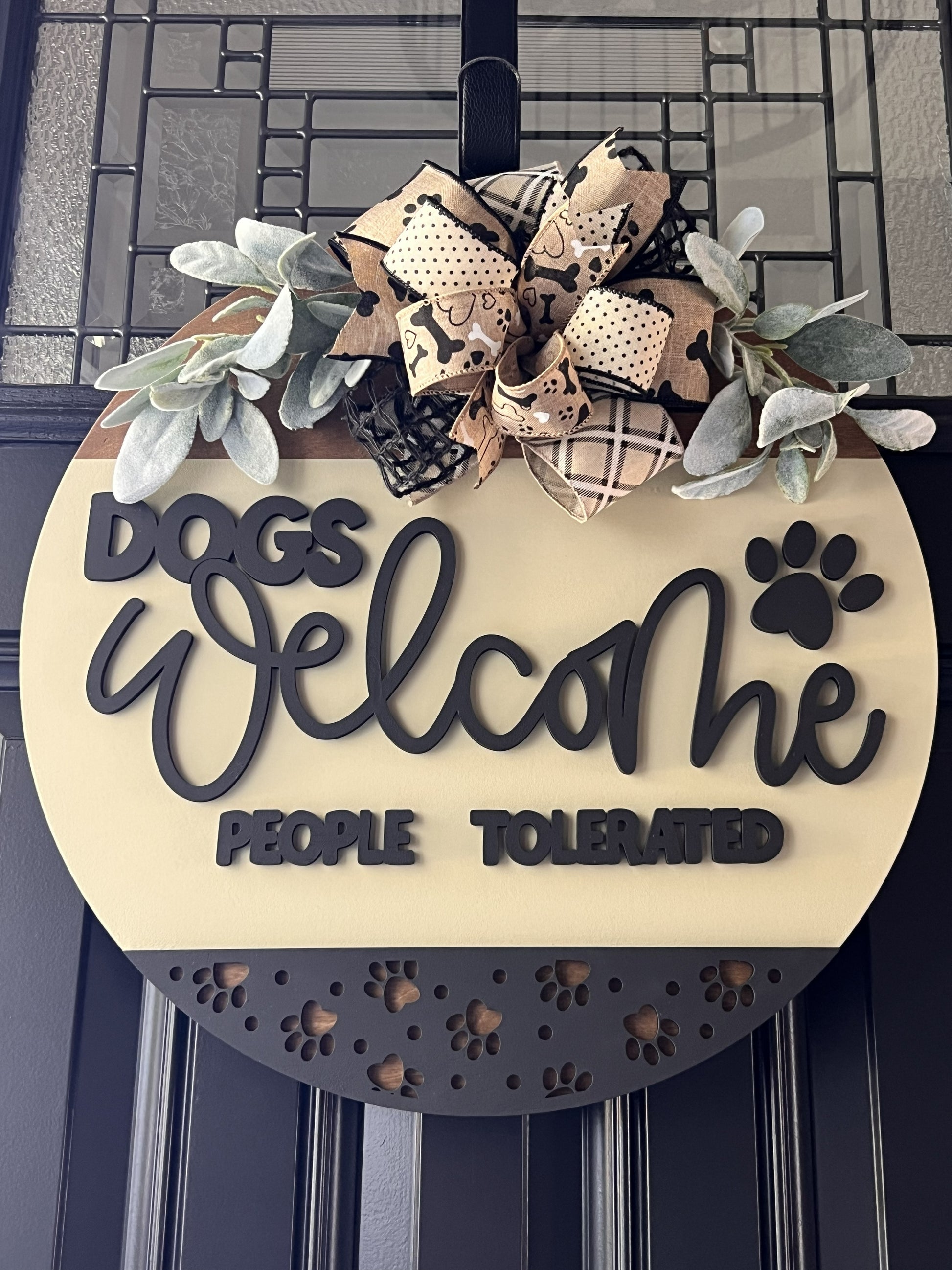 “Dogs Welcome People Tolerated wooden door hanger with layered lettering, paw print accents and decorative bow on front door”