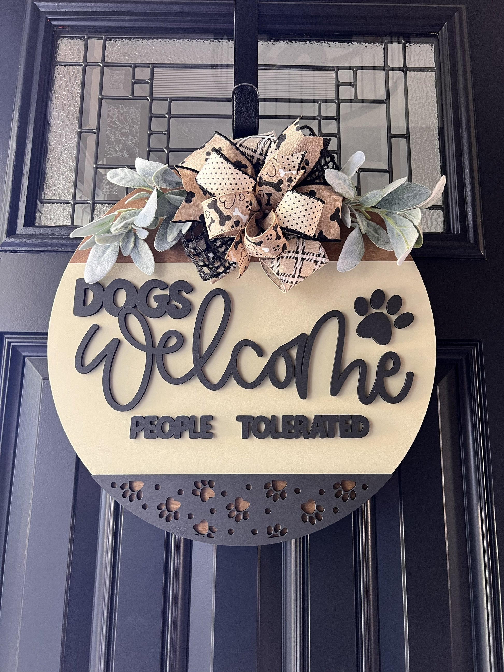 “Dogs Welcome People Tolerated wooden door hanger with layered lettering, paw print accents and decorative bow on front door”