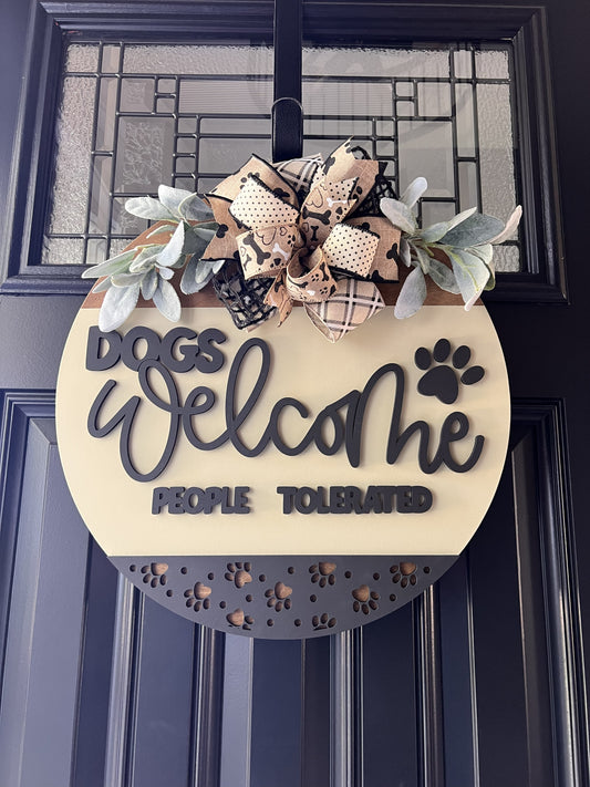 “Dogs Welcome People Tolerated wooden door hanger with layered lettering, paw print accents and decorative bow on front door”