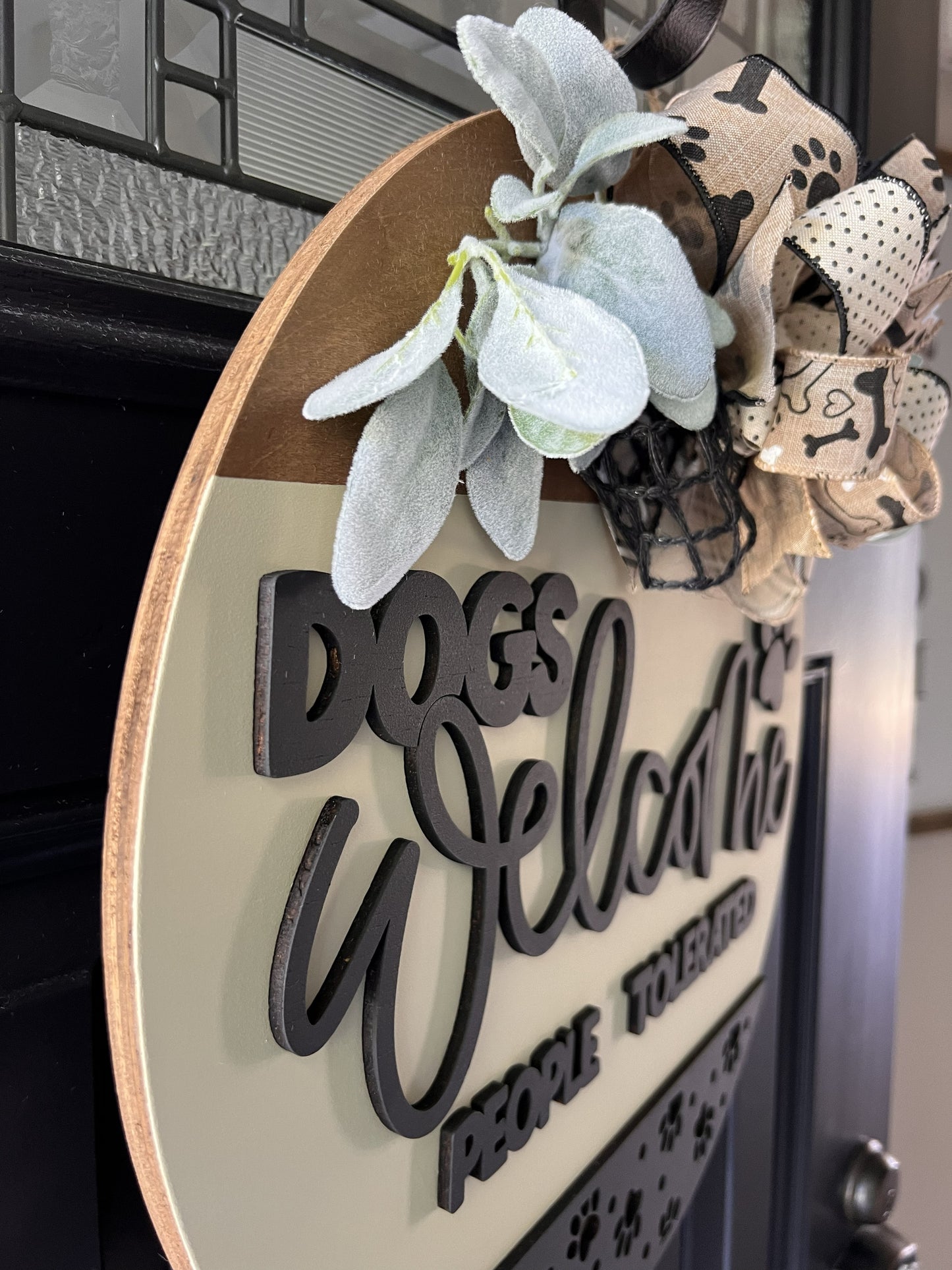 “Dogs Welcome People Tolerated wooden door hanger with layered lettering, paw print accents and decorative bow on front door”