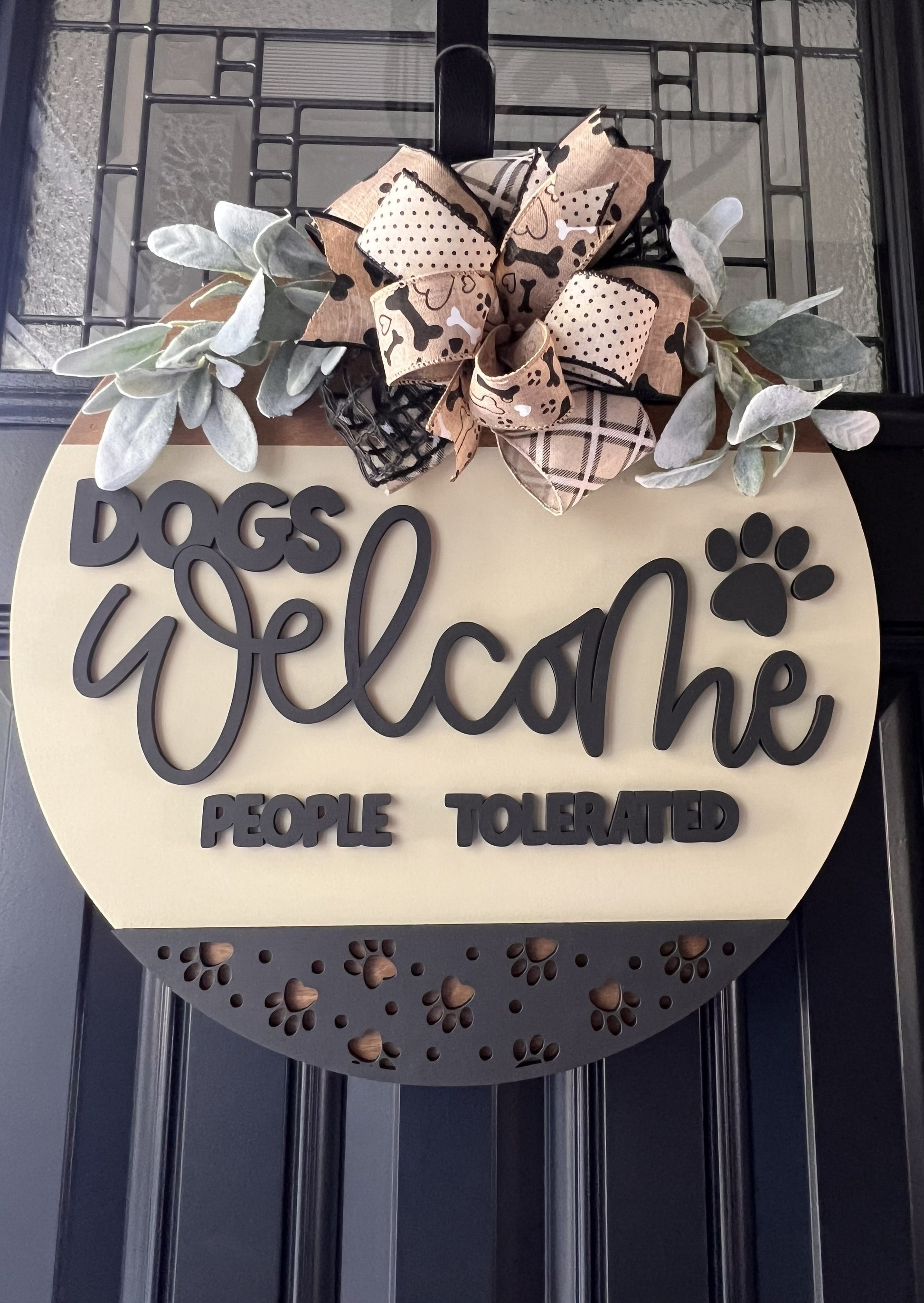 “Dogs Welcome People Tolerated wooden door hanger with layered lettering, paw print accents and decorative bow on front door”