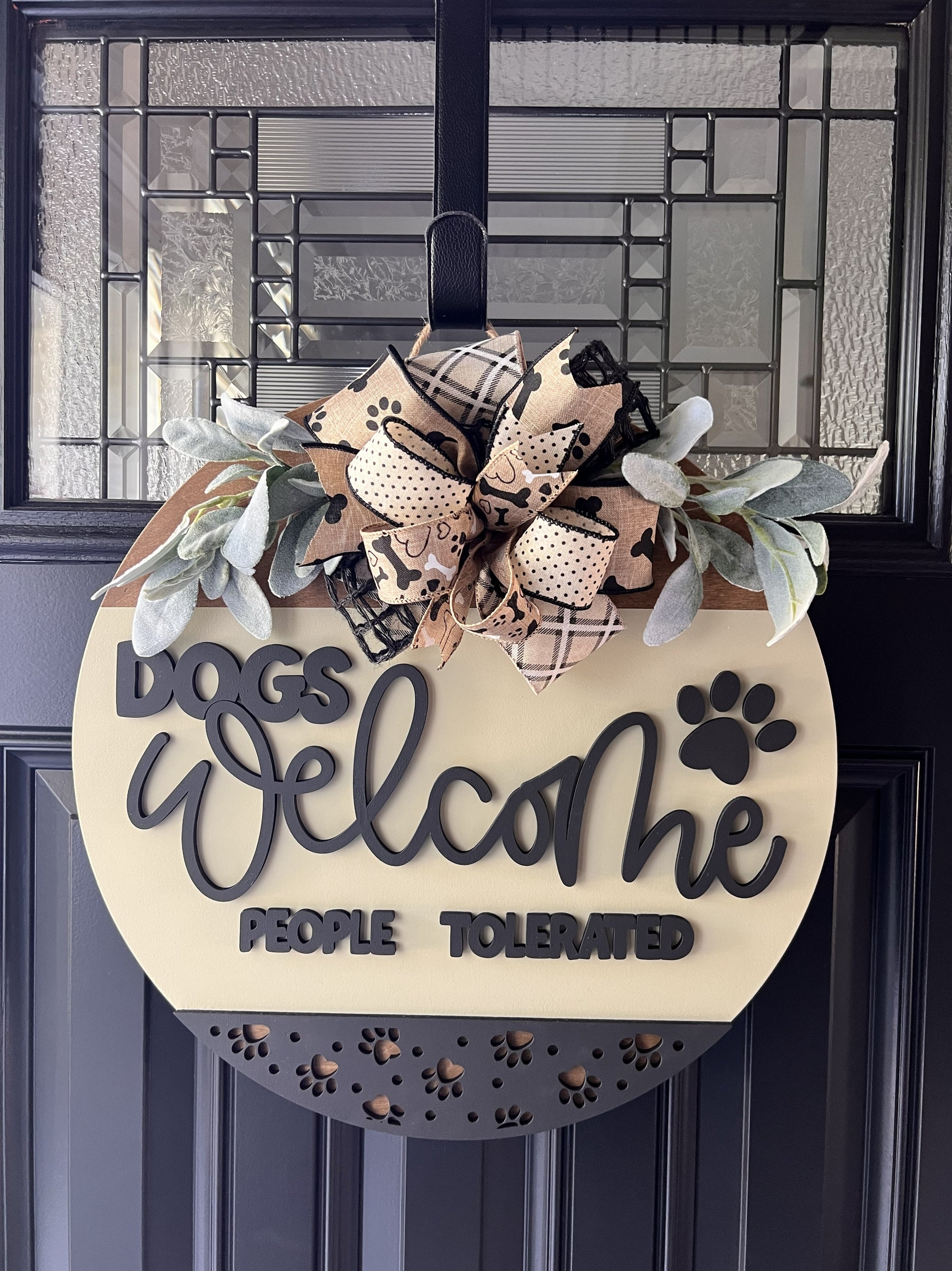 “Dogs Welcome People Tolerated wooden door hanger with layered lettering, paw print accents and decorative bow on front door”