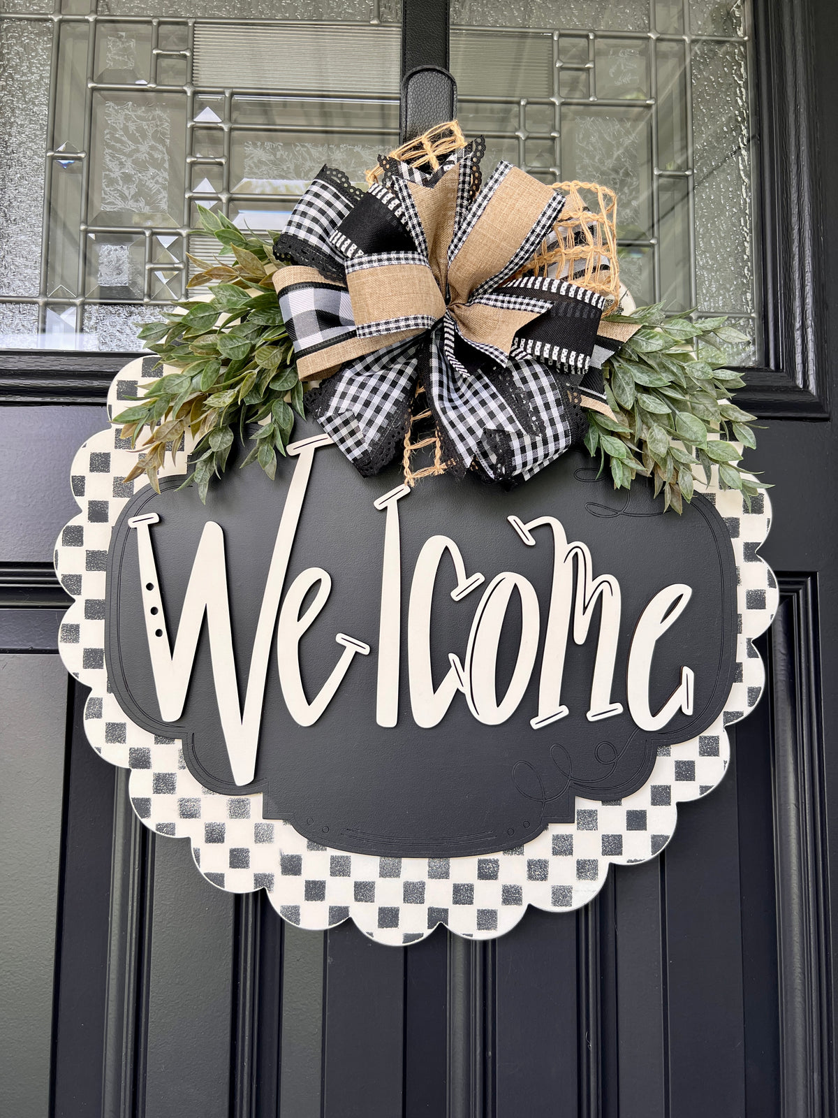 Close-up of black and white welcome door hanger with buffalo check, burlap, and jute ribbon bow and faux greenery