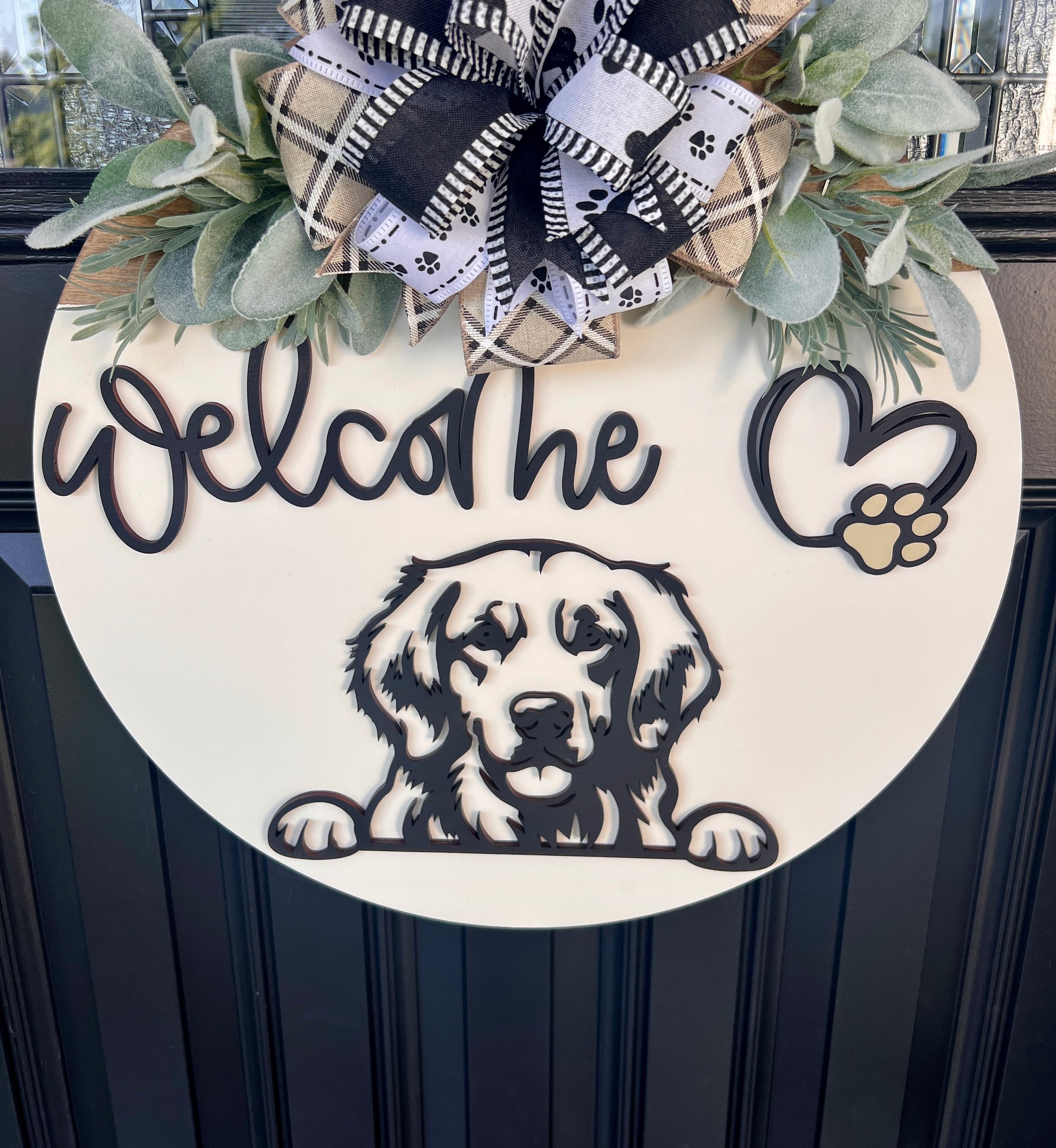 A round white wooden door wreath featuring a black silhouette of a golden retriever dog, the word 'Welcome' in script, a heart outline with a paw print, and a large black-and-cream plaid ribbon bow with green foliage at the top.