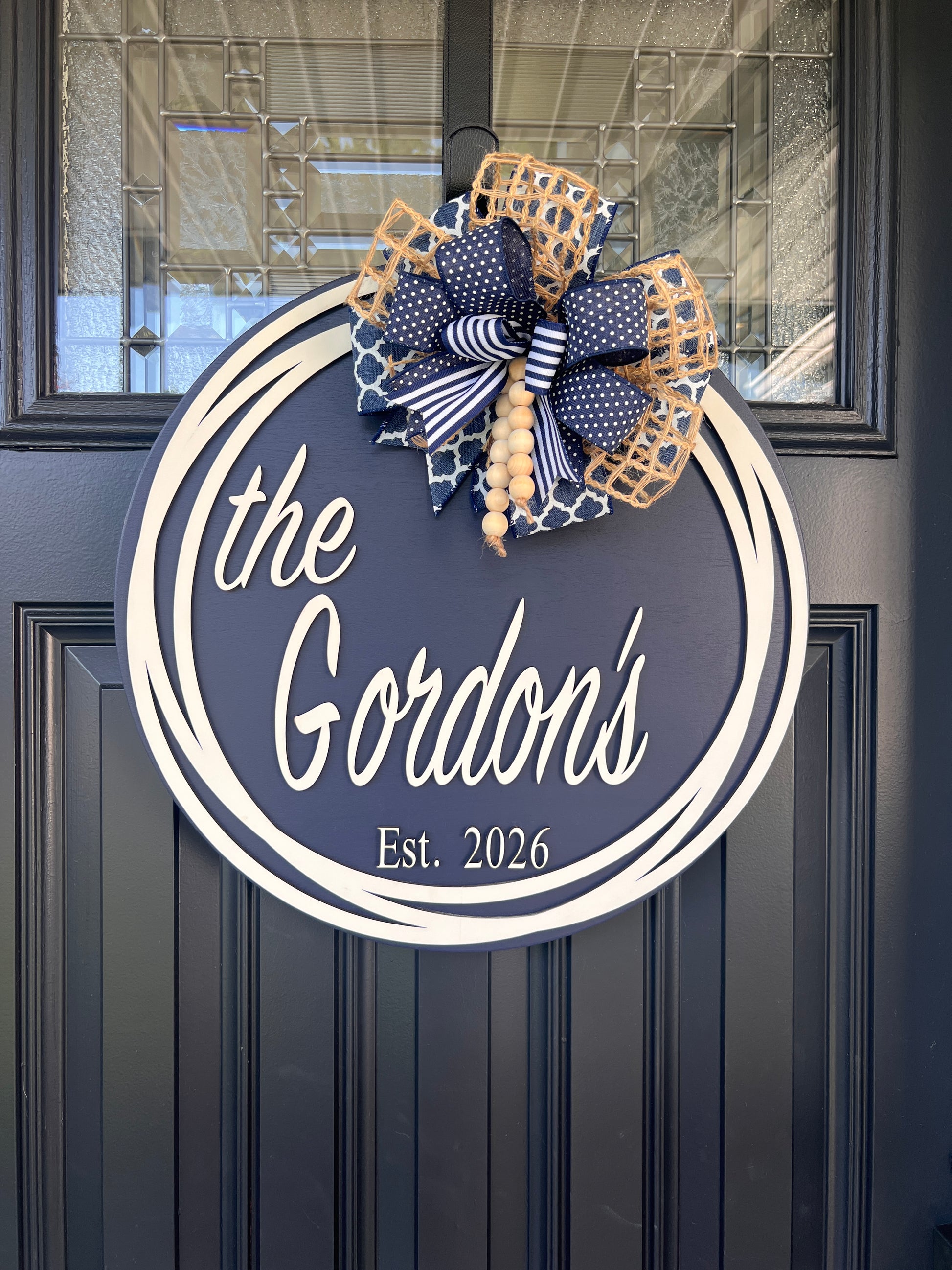 A personalized round door sign mounted on a dark paneled front door. The sign is navy blue with white script text  with family name and est. date 
