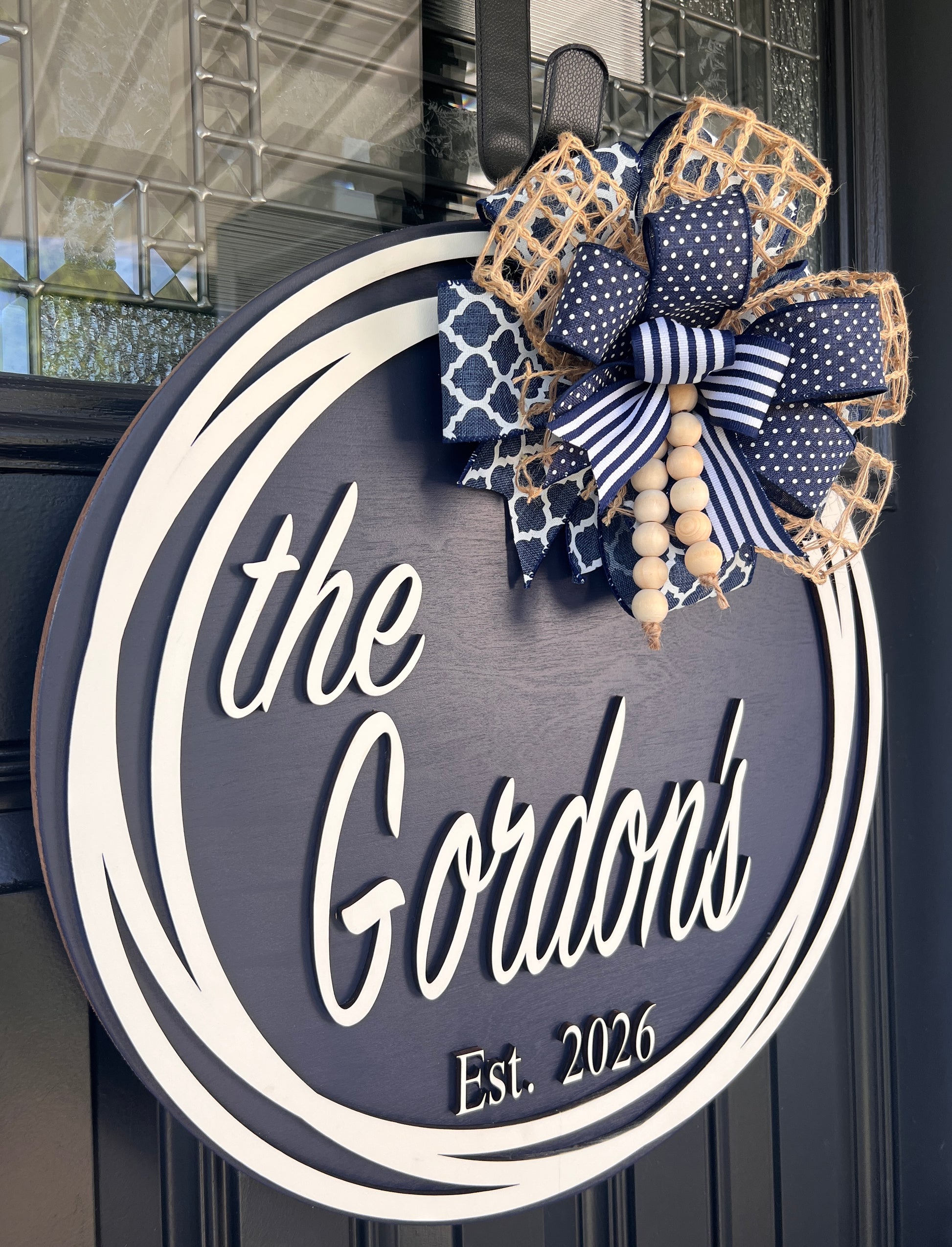 A personalized round wooden door sign reading 'the Gordon's Est. 2026' in white script, decorated with a navy and white patterned bow, beige lattice ribbon, and wooden beads, hanging on a dark paneled door.