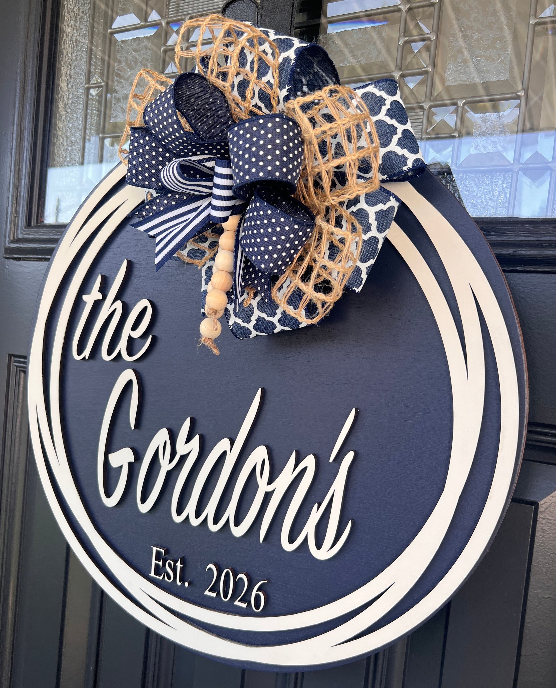 Custom name door hanger angled close up showing the Gordons script lettering and bow detail with navy polka dot stripe and jute burlap ribbons