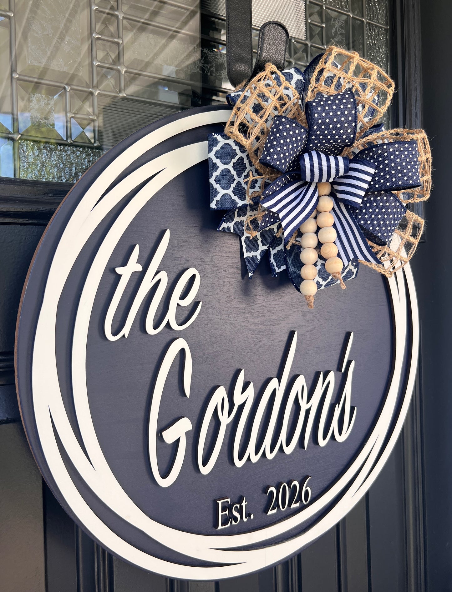 Custom family name door hanger full view on black front door showing complete navy round with cream double circle frame and wood bead bow accent