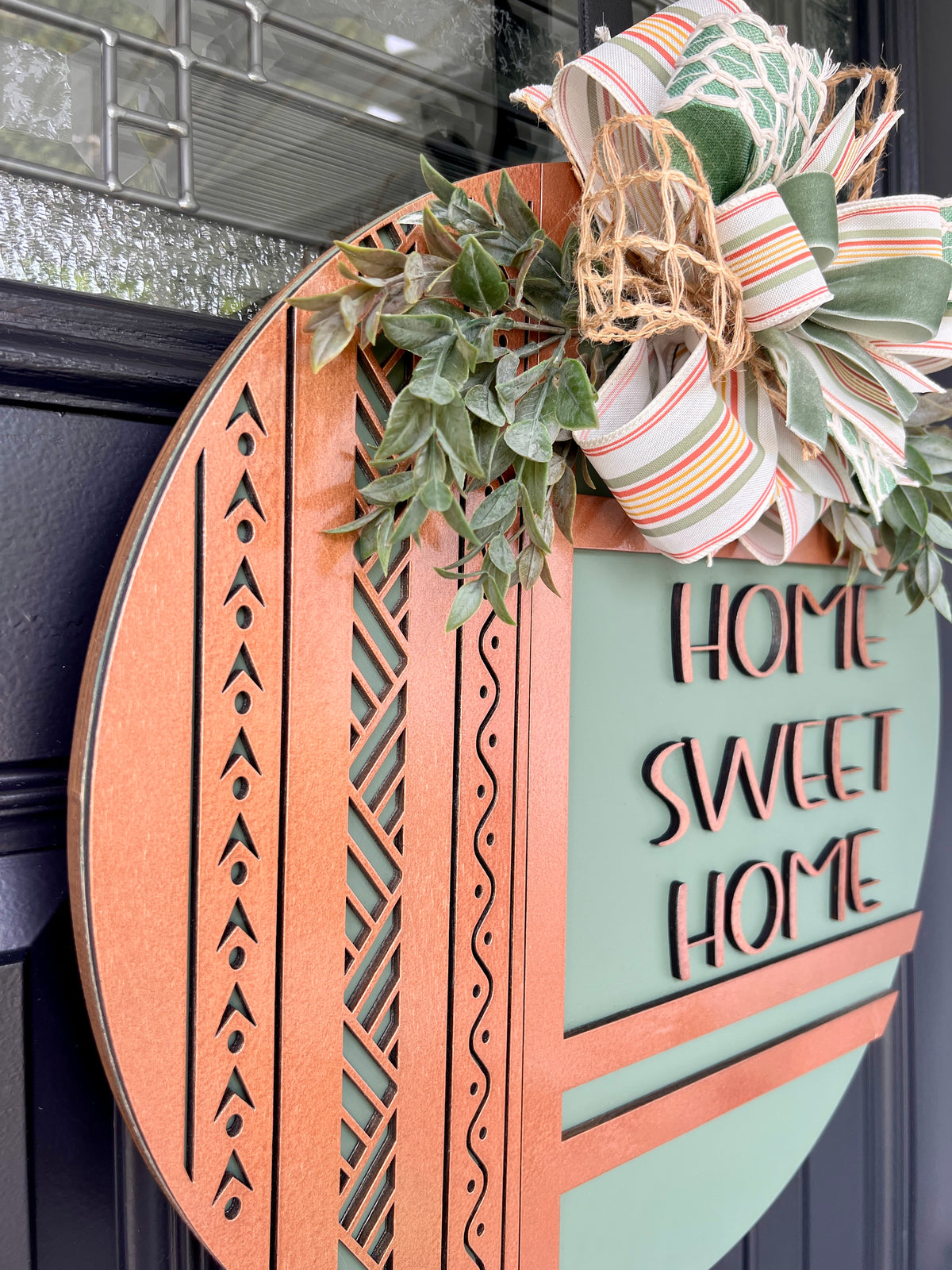 Angled close-up of raised copper Home Sweet Home lettering on a sage green birchwood front door sign with greenery and bow