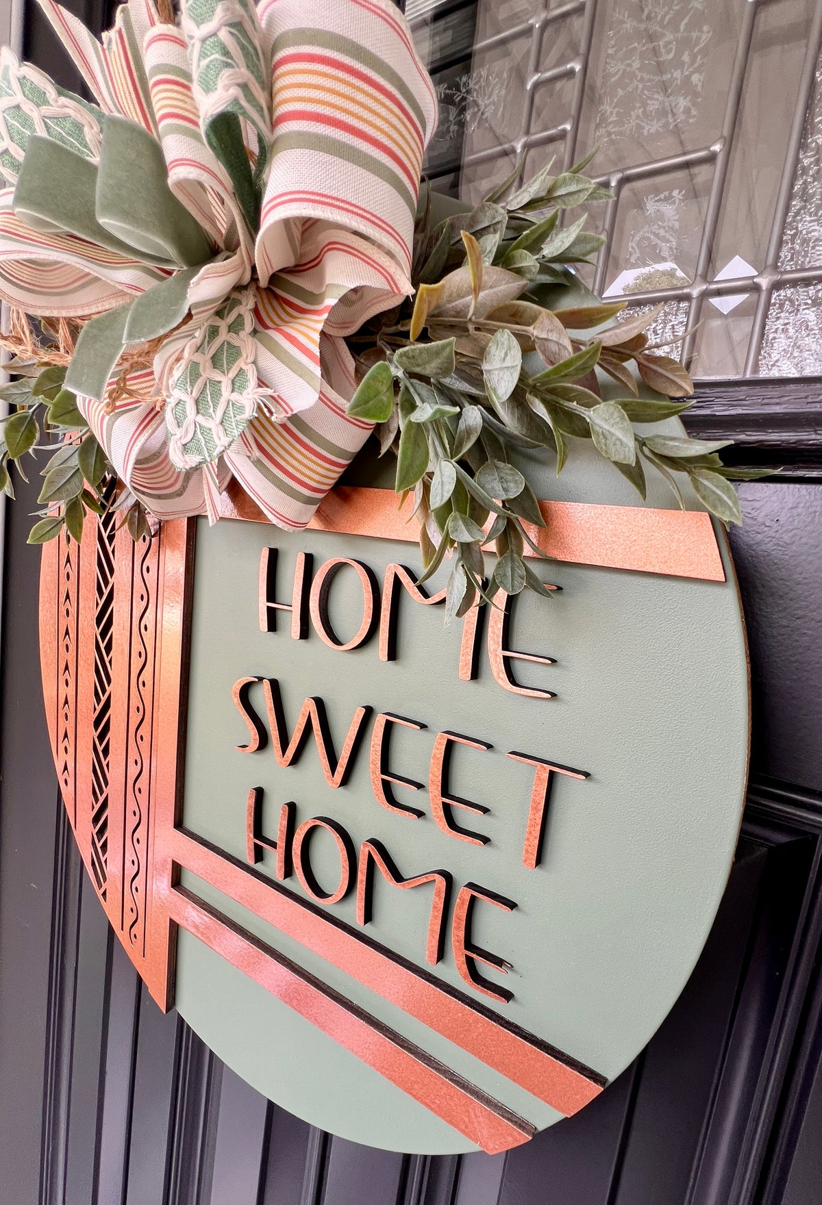 Close-up of layered copper wood detailing and geometric laser-cut patterns on a sage green Home Sweet Home front door sign