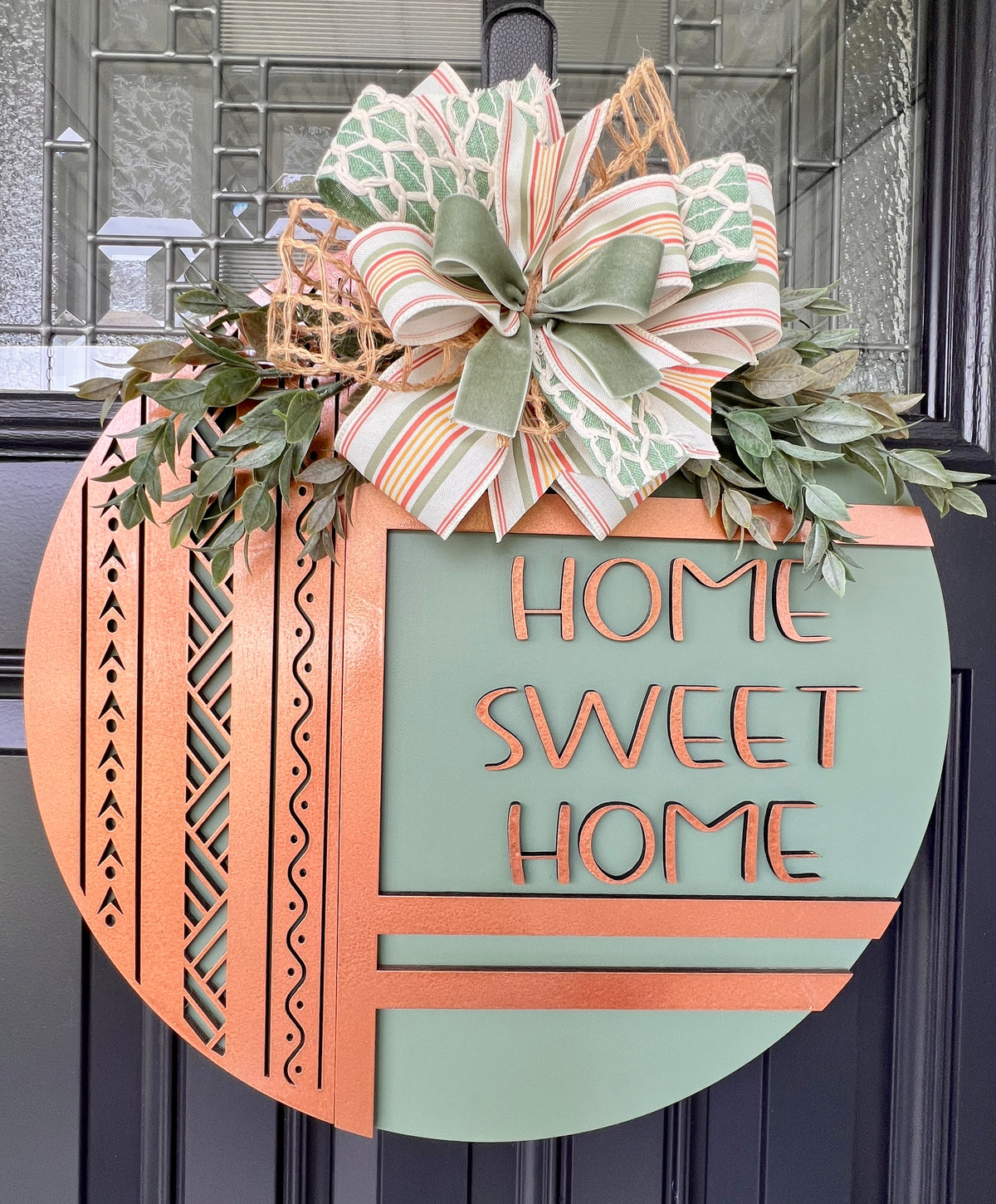 Sage green and copper Home Sweet Home front door hanger displayed on a black door with greenery and a hand-tied multi-ribbon bow