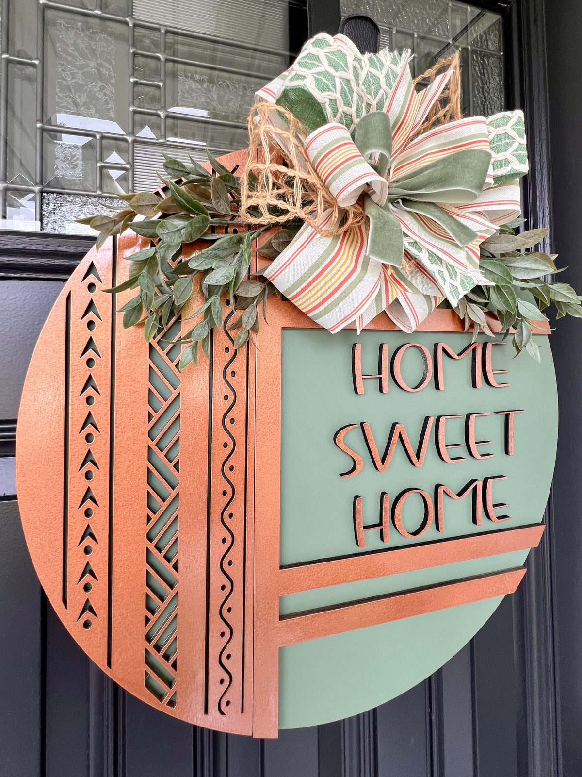 Handmade round sage green farmhouse door sign with copper Home Sweet Home lettering, geometric cutouts, greenery, and decorative bow