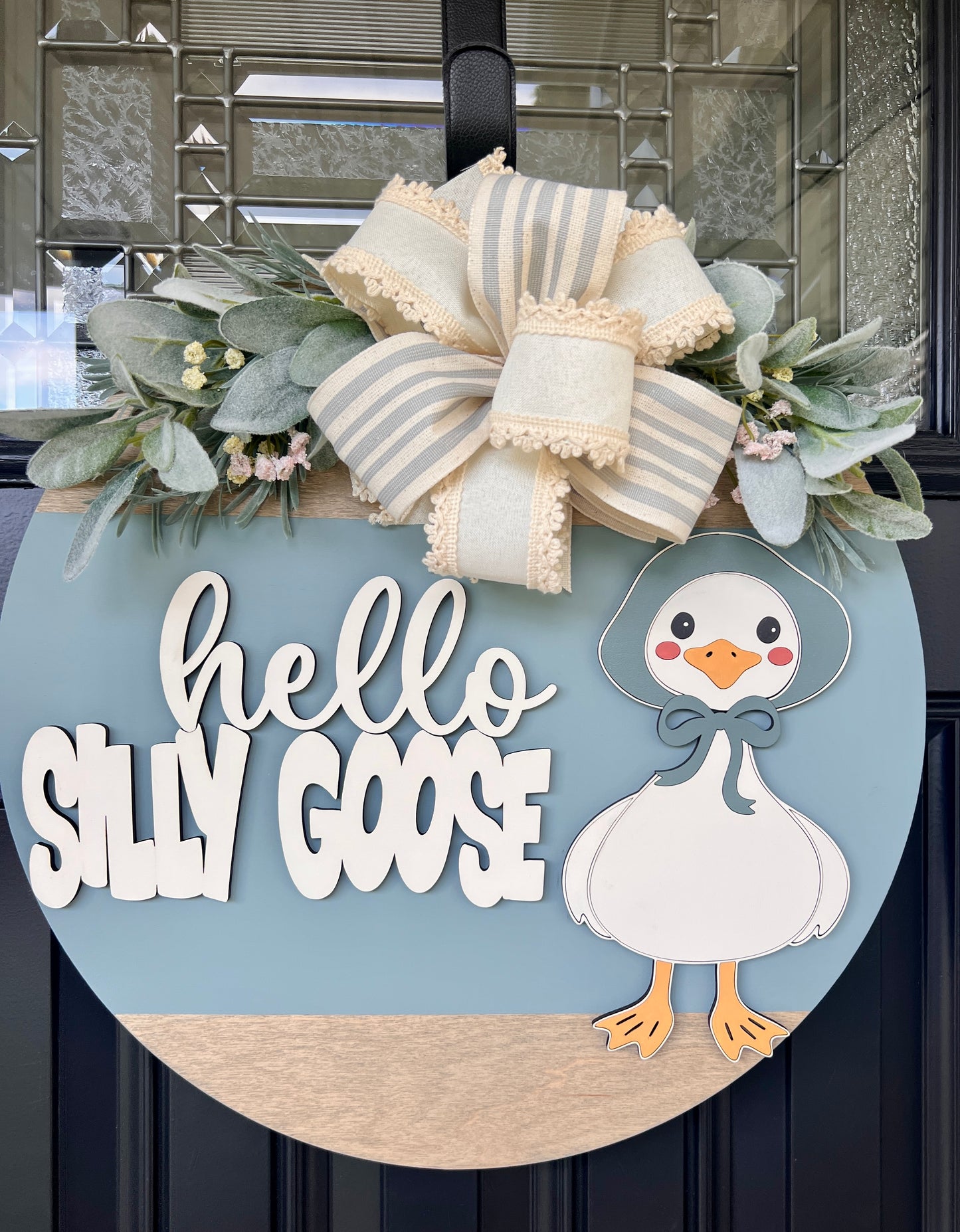 Handmade Hello Silly Goose door hanger with layered lettering, blue background, farmhouse goose illustration, greenery, and designer bow on a black front door.