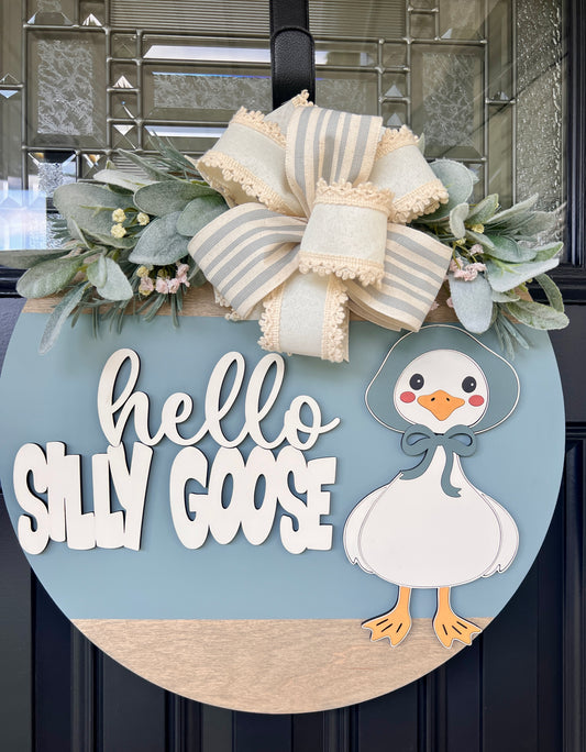 Handmade Hello Silly Goose door hanger with layered lettering, blue background, farmhouse goose illustration, greenery, and designer bow on a black front door.