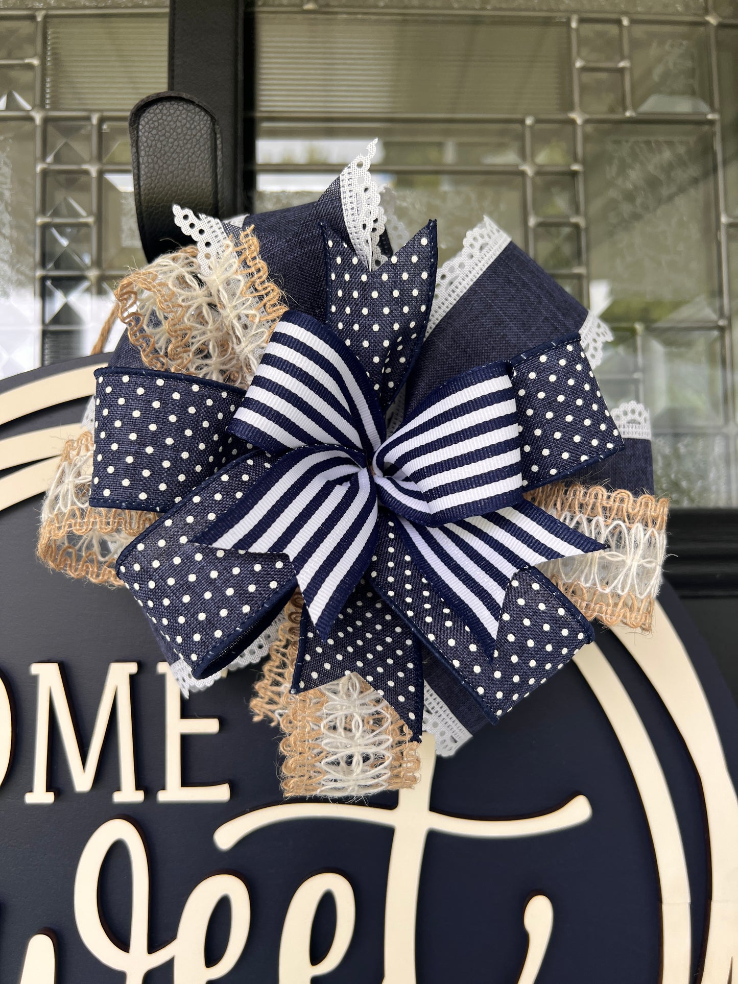 A decorative autumn wreath with a large navy and white striped and polka-dot bow, layered with beige burlap and white lace, hanging on a dark navy sign that reads 'Home Sweet Home'.