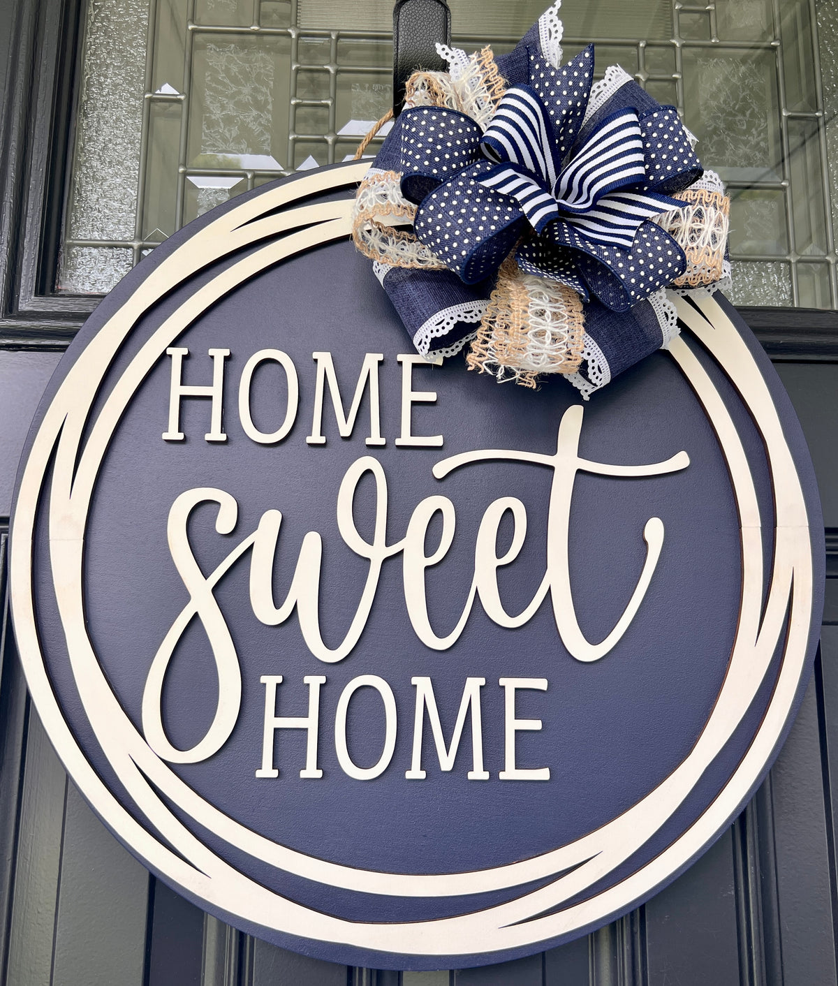 A round navy blue and white wooden door sign with the words 'HOME sweet HOME' in white lettering, decorated with a large navy, white, and beige burlap ribbon bow at the top, shown hanging on a dark paneled door.
