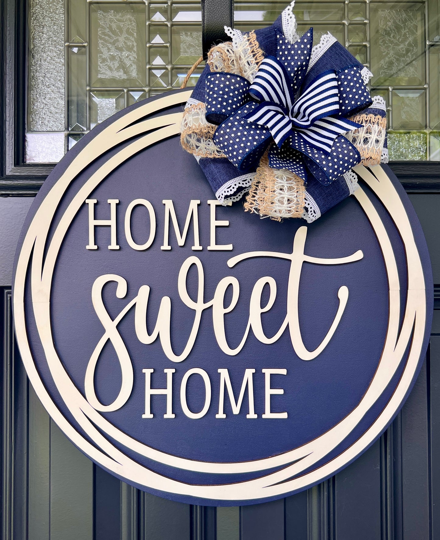 A round navy blue wooden door wreath with the phrase 'HOME sweet HOME' in white lettering, decorated with a large navy, white, and beige ribbon bow at the top, hanging on a dark paneled front door.