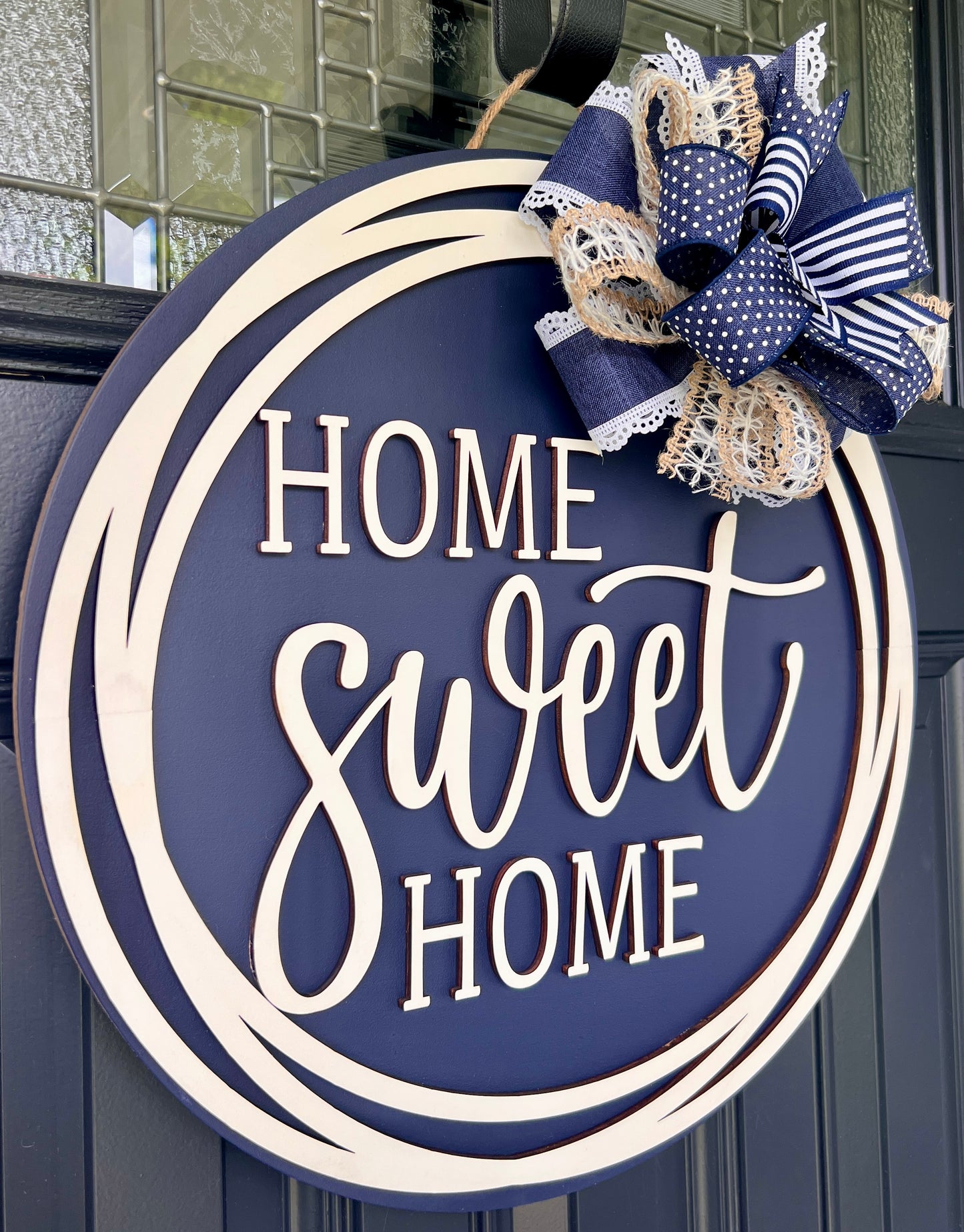 Round navy blue and cream wooden door sign reading “Home Sweet Home,” decorated with a navy, white, and beige bow on a dark paneled door.