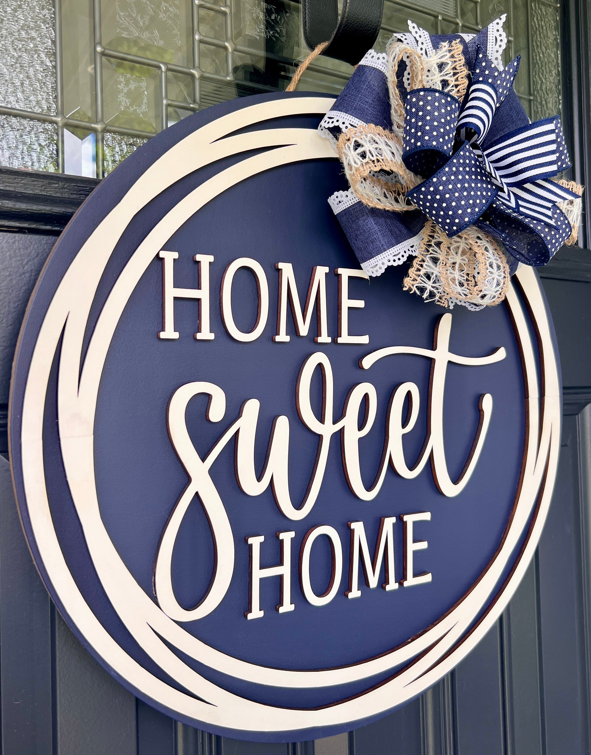 Round navy blue and cream wooden door sign reading “Home Sweet Home,” decorated with a navy, white, and beige bow on a dark paneled door.