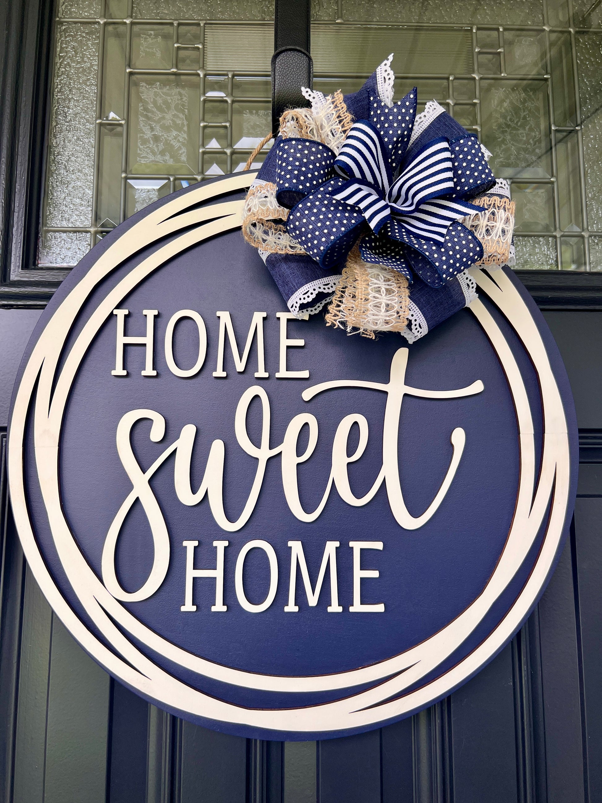 A round navy blue and beige wooden wreath hanging on a dark front door, featuring the phrase “HOME sweet HOME” in white lettering and topped with a decorative ribbon bow in navy, white, and tan.