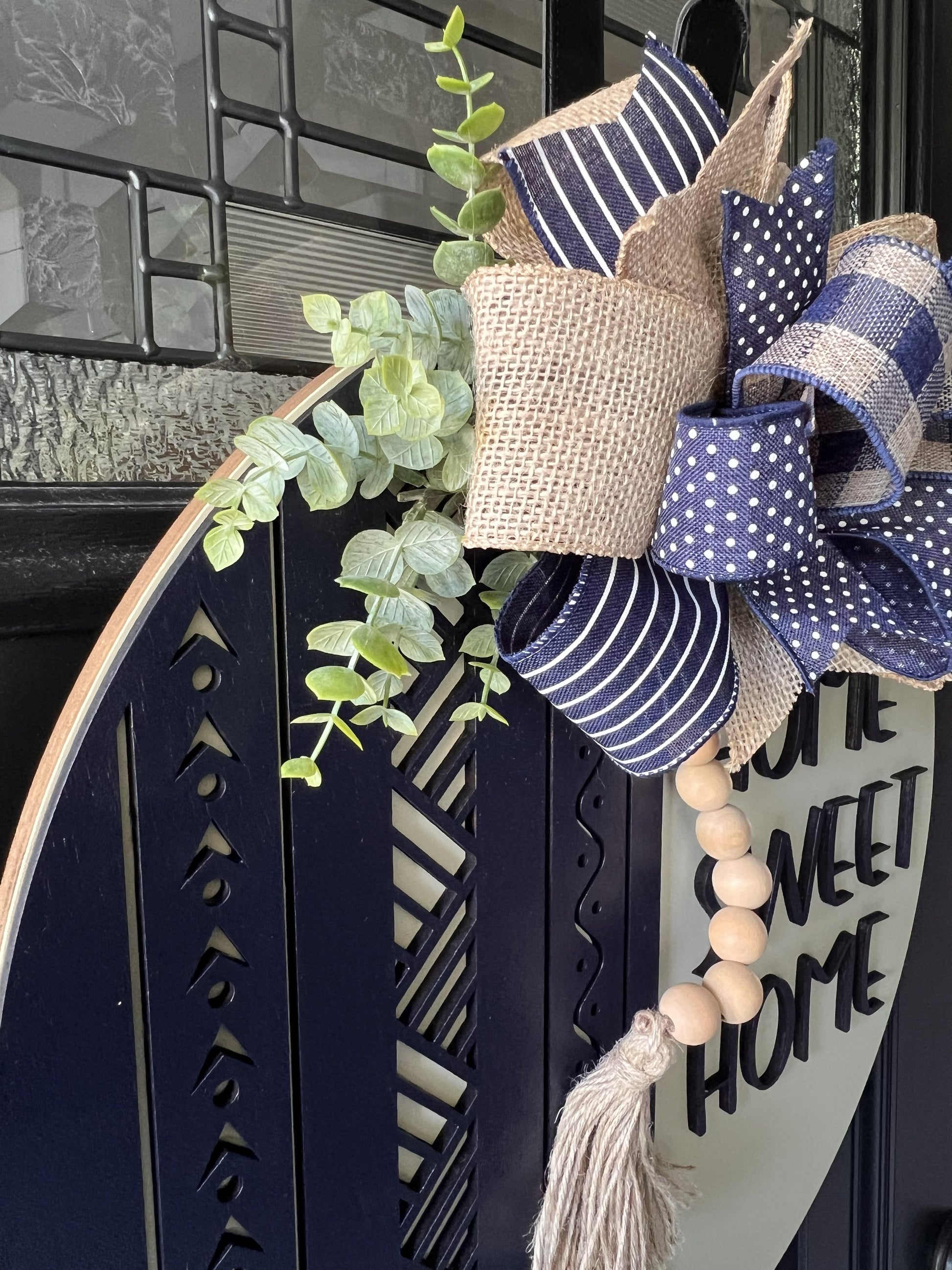 Decorative wreath with blue and beige ribbons, eucalyptus leaves, and a wooden beaded tassel on a door.