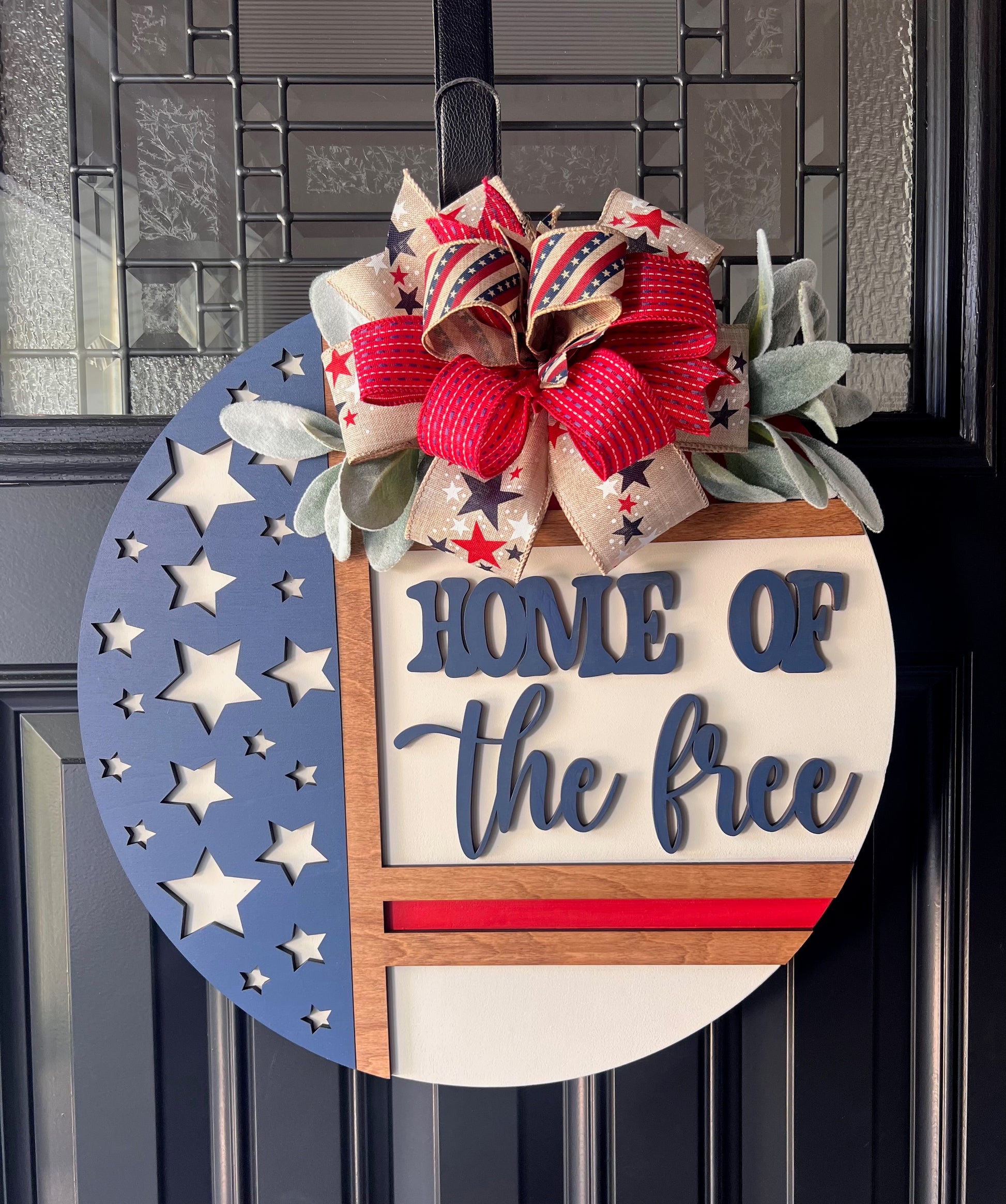 Home of the Free patriotic door hanger - navy and cream version with laser-cut star field left panel, 3D navy lettering on cream right panel with red stripe accent, layered patriotic star and stripe ribbon bow with lambs ear greenery on black front door