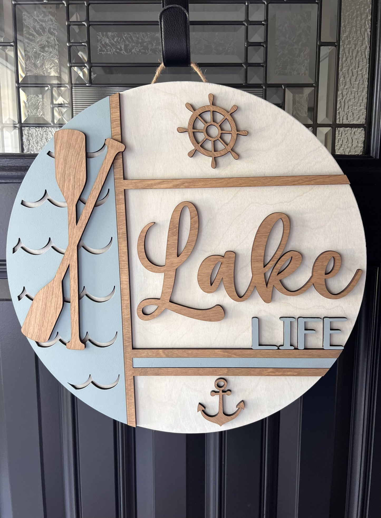 Decorative sign with 'Lake Life' text, oar, wheel, and anchor on a light blue and white background.