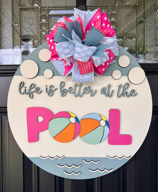 Handcrafted round door hanger featuring the phrase "Life Is Better at the Pool" with colorful beach ball accents, summer colors, and a bright pink and blue designer bow displayed on a front door.