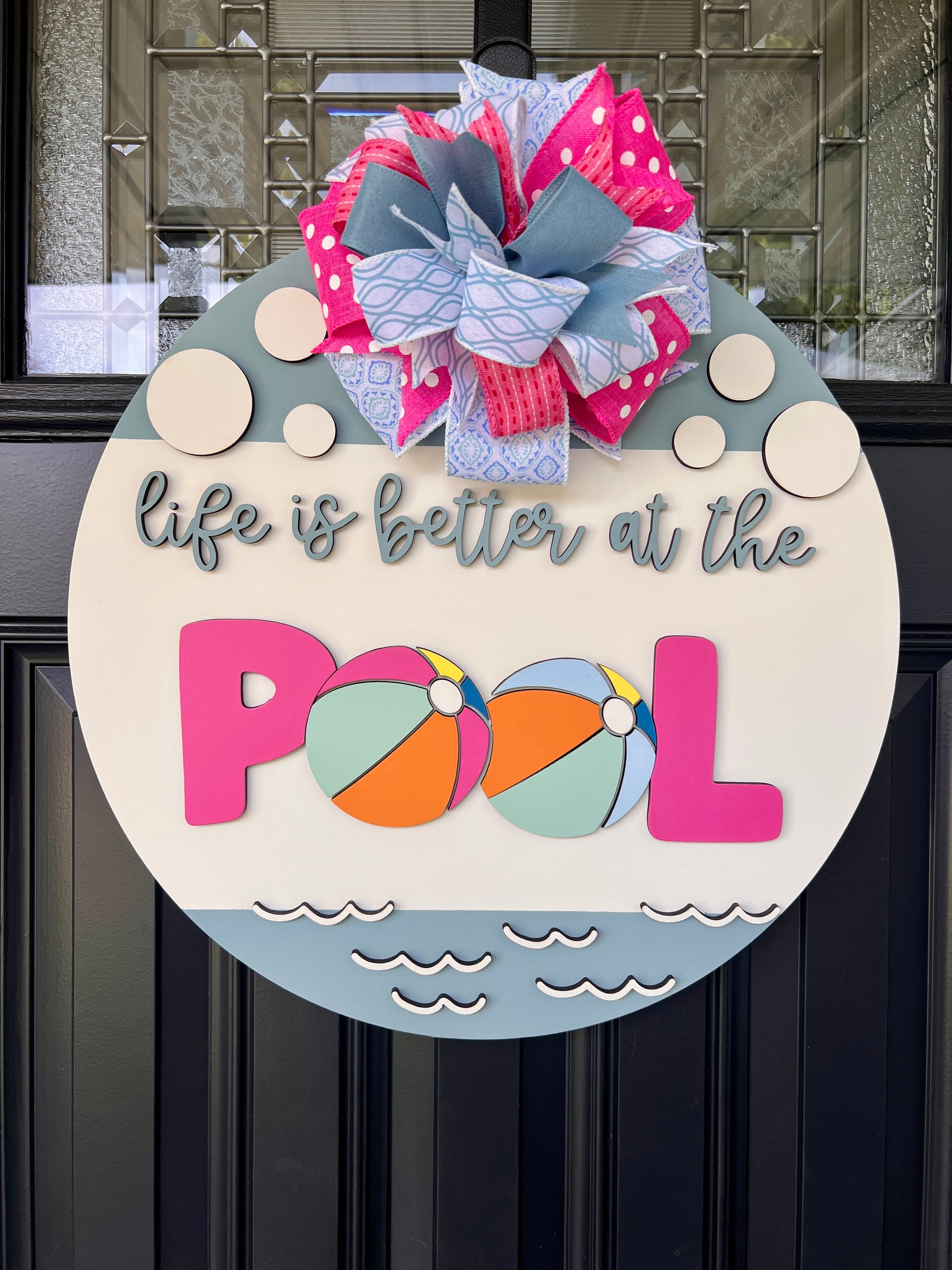 Handcrafted round door hanger featuring the phrase "Life Is Better at the Pool" with colorful beach ball accents, summer colors, and a bright pink and blue designer bow displayed on a front door.