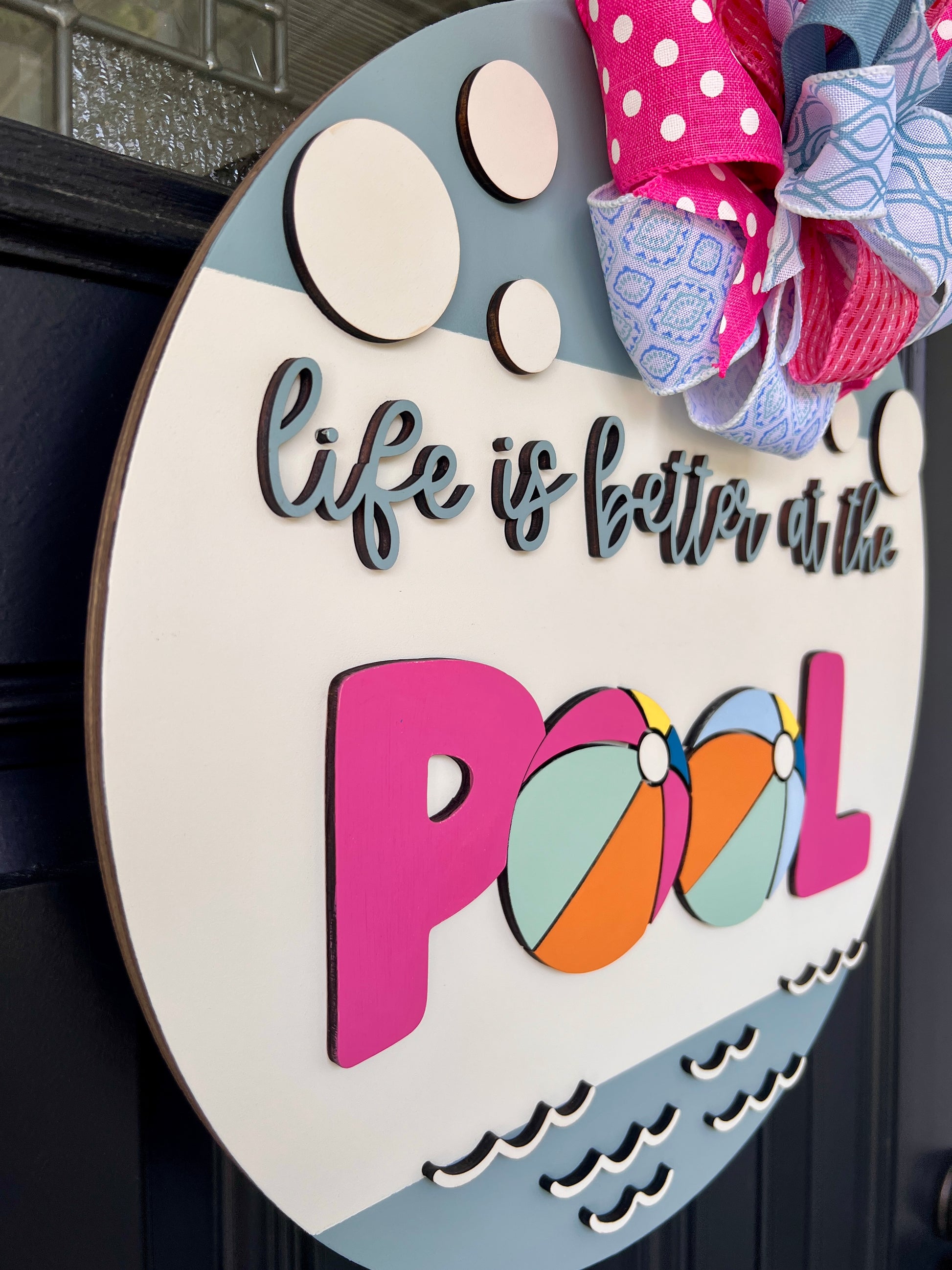 Handcrafted round door hanger featuring the phrase "Life Is Better at the Pool" with colorful beach ball accents, summer colors, and a bright pink and blue designer bow displayed on a front door.