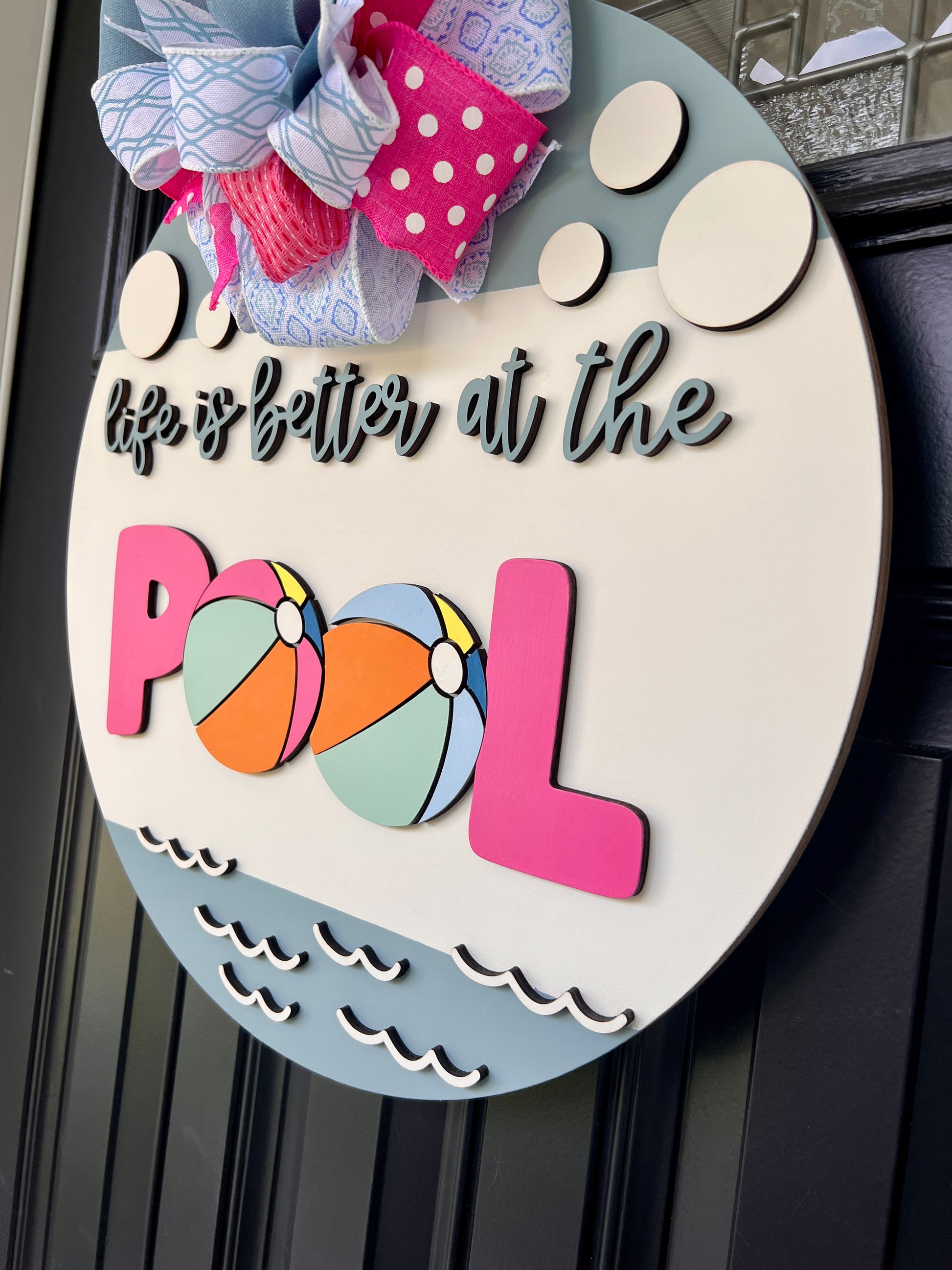 Handcrafted round door hanger featuring the phrase "Life Is Better at the Pool" with colorful beach ball accents, summer colors, and a bright pink and blue designer bow displayed on a front door.