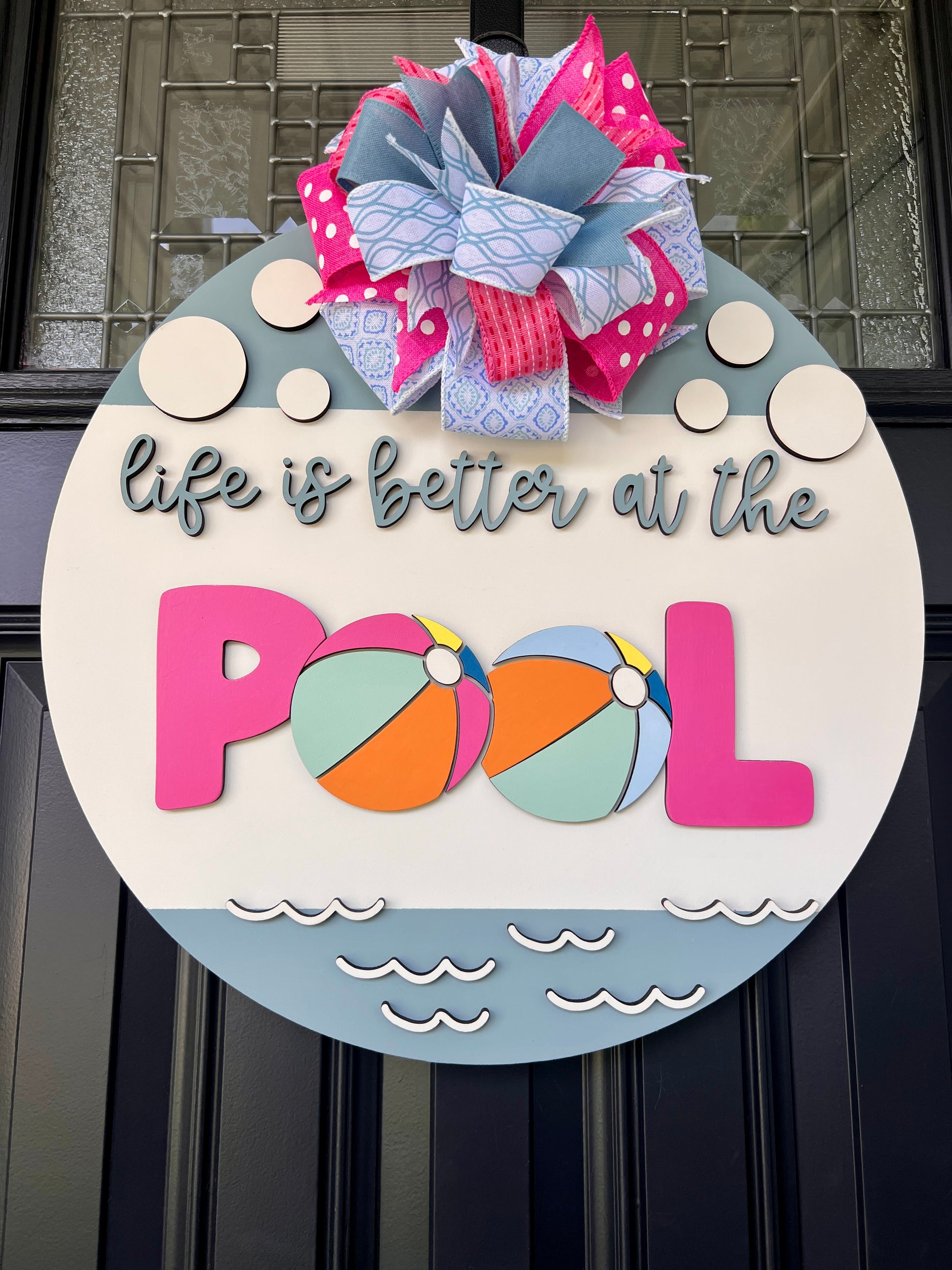 Handcrafted round door hanger featuring the phrase "Life Is Better at the Pool" with colorful beach ball accents, summer colors, and a bright pink and blue designer bow displayed on a front door.