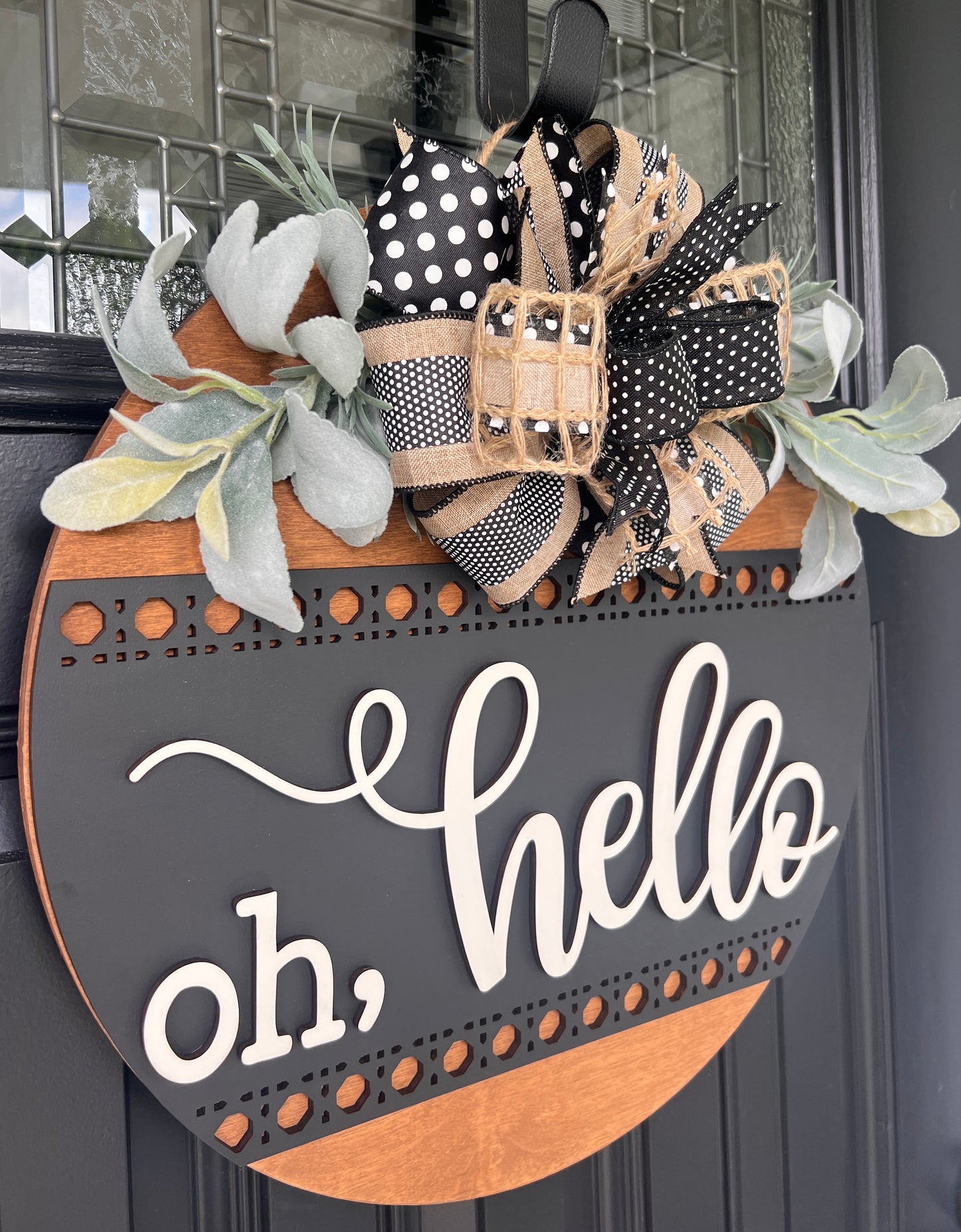 A rustic round wooden door wreath hanging on a dark front door. The wreath has a black top section with white script reading 'oh, hello', a natural wood bottom section, and a large decorative bow made of black-and-white polka dot ribbon, burlap, and black ribbon with white dots.