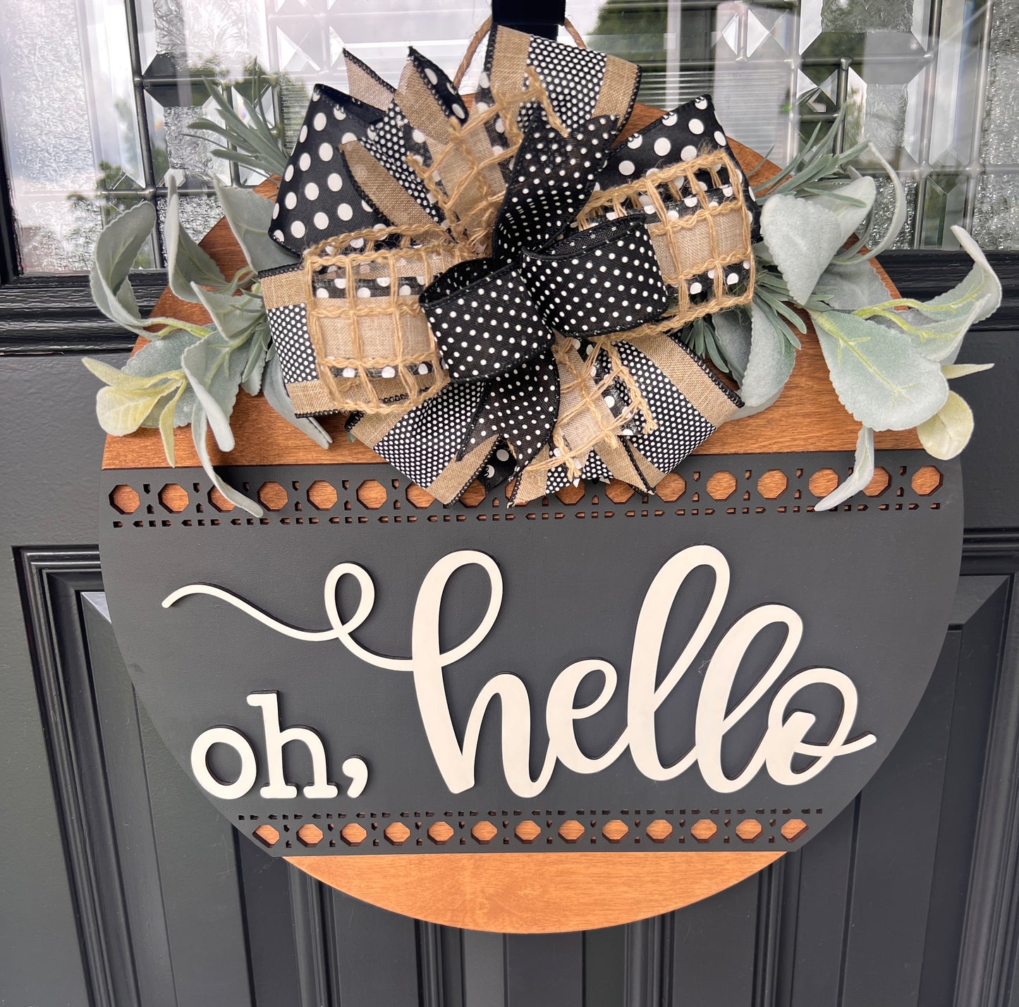 A decorative wreath hanging on a dark paneled door. The wreath has a large bow made of black-and-white polka dot ribbon, tan burlap, and patterned ribbon, with green eucalyptus leaves.  Layered script "Oh Hello"