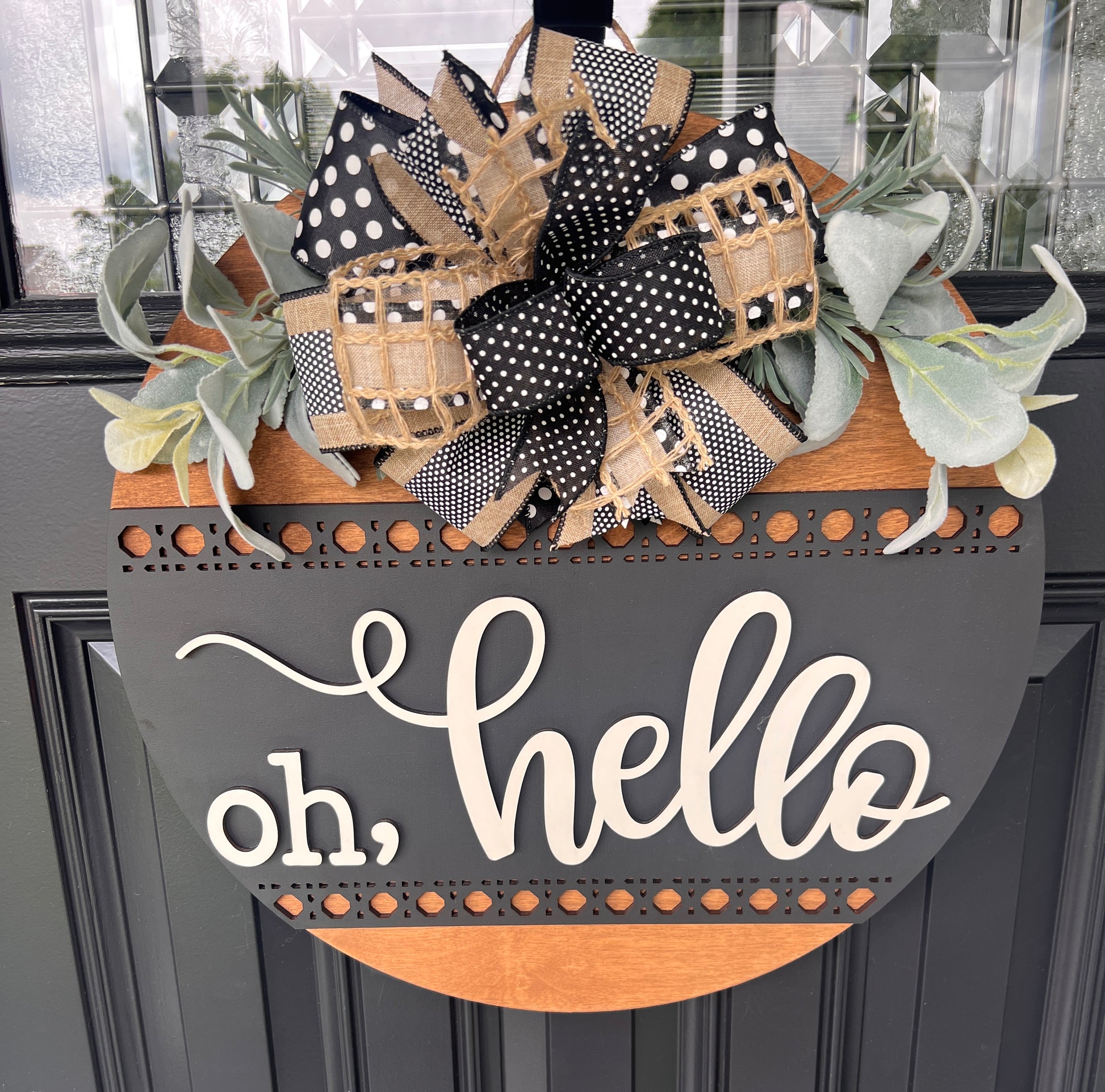 A decorative wreath hanging on a dark paneled door. The wreath has a large bow made of black-and-white polka dot ribbon, tan burlap, and patterned ribbon, with green eucalyptus leaves.  Layered script "Oh Hello"