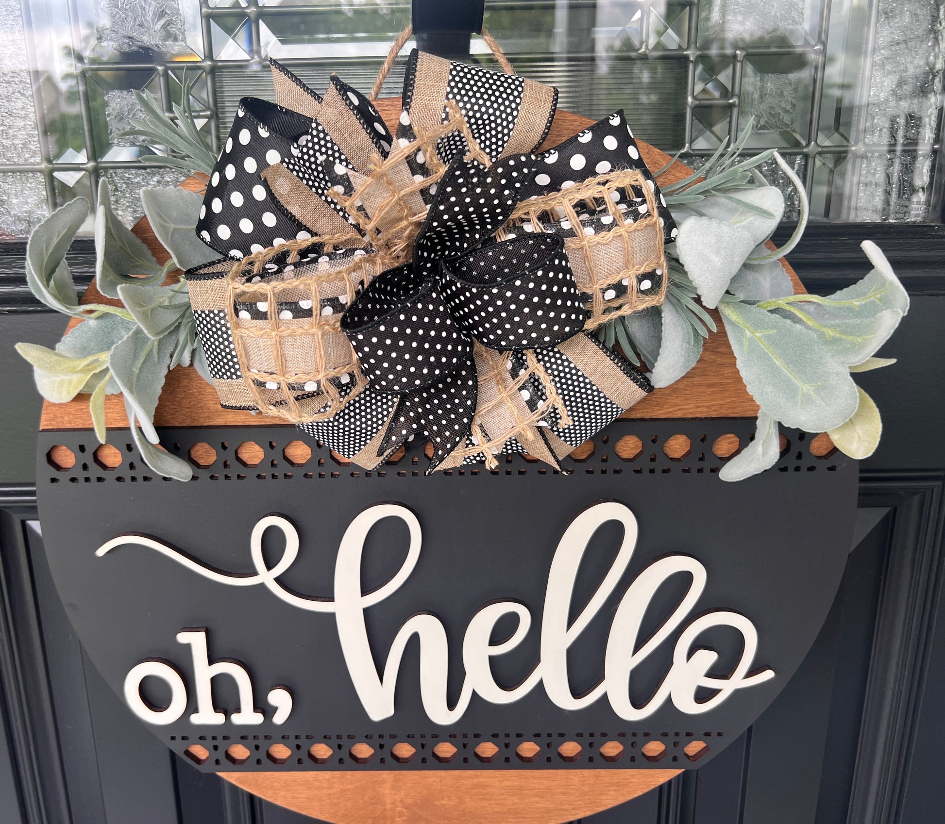 A decorative black and wood door sign with the phrase "oh, hello" in white script, styled with a large black-and-tan polka-dot and burlap bow and faux eucalyptus leaves in front of a glass-paneled door.