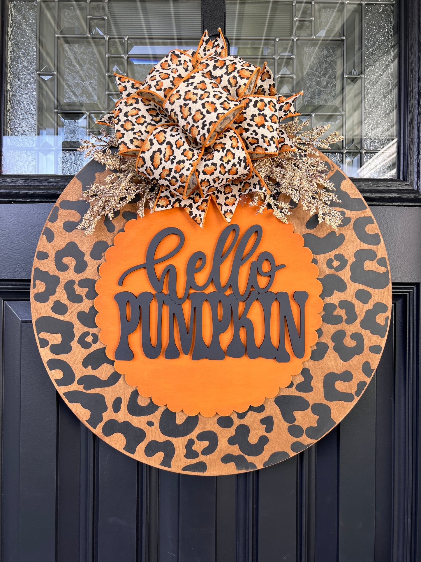 Hello Pumpkin leopard print fall door hanger with layered wood lettering, designer bow, seasonal greenery, and woodgrain animal print background displayed on a front door